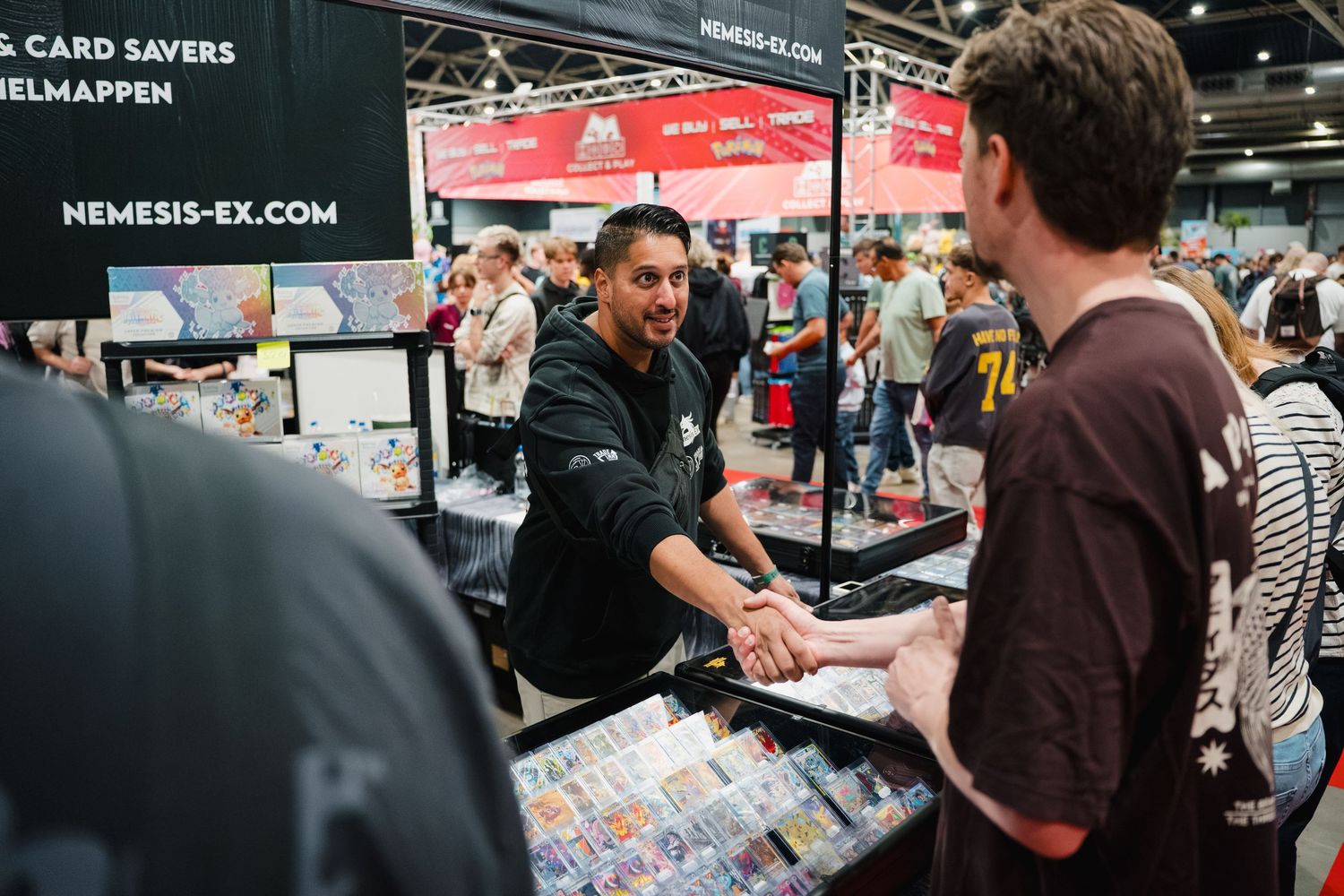 Vendor at Nemesis-EX trading card booth shakes hands with customer at busy gaming convention.