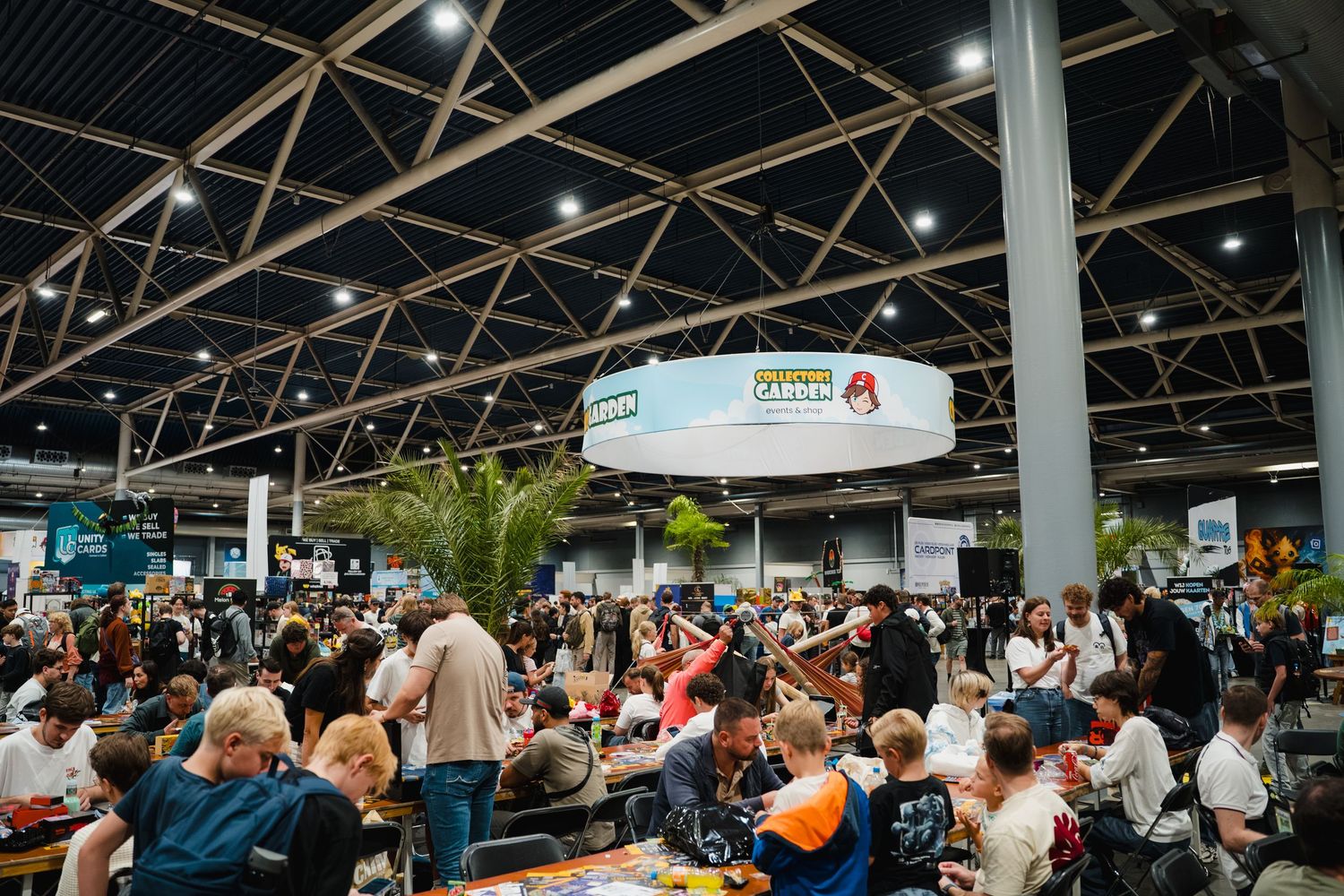 Crowded convention hall with vendors, attendees, and a large circular hanging sign in a spacious industrial venue.