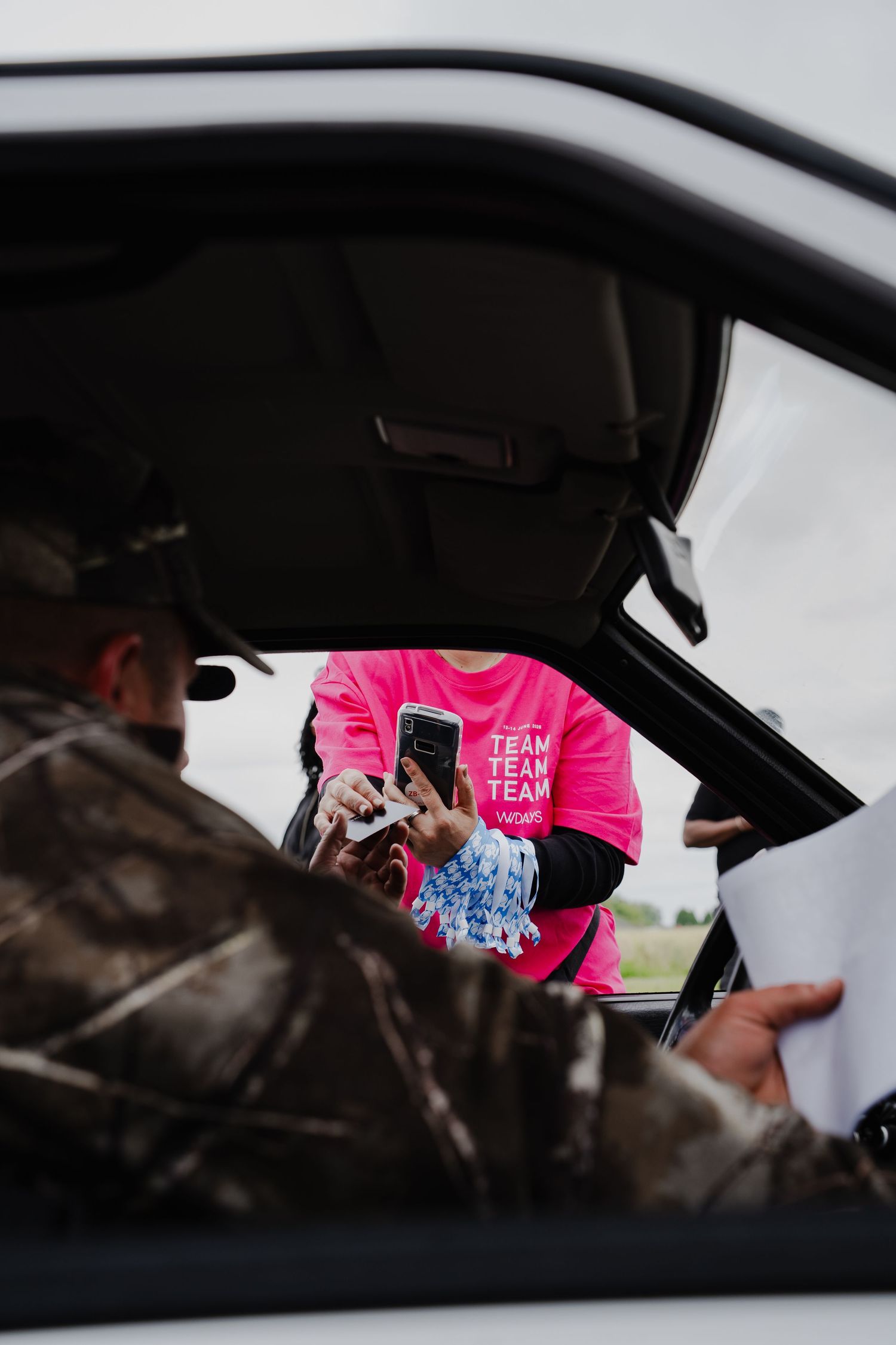 Person in pink team shirt uses phone at car window while man in camo jacket sits inside white vehicle.
