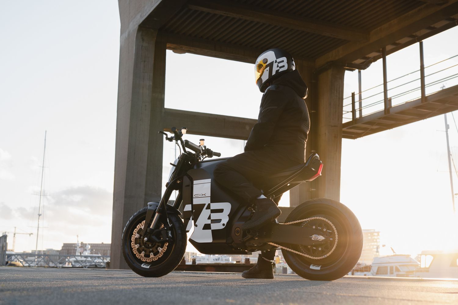 Motorcycle silhouetted against bright sunset with industrial bridge in background.