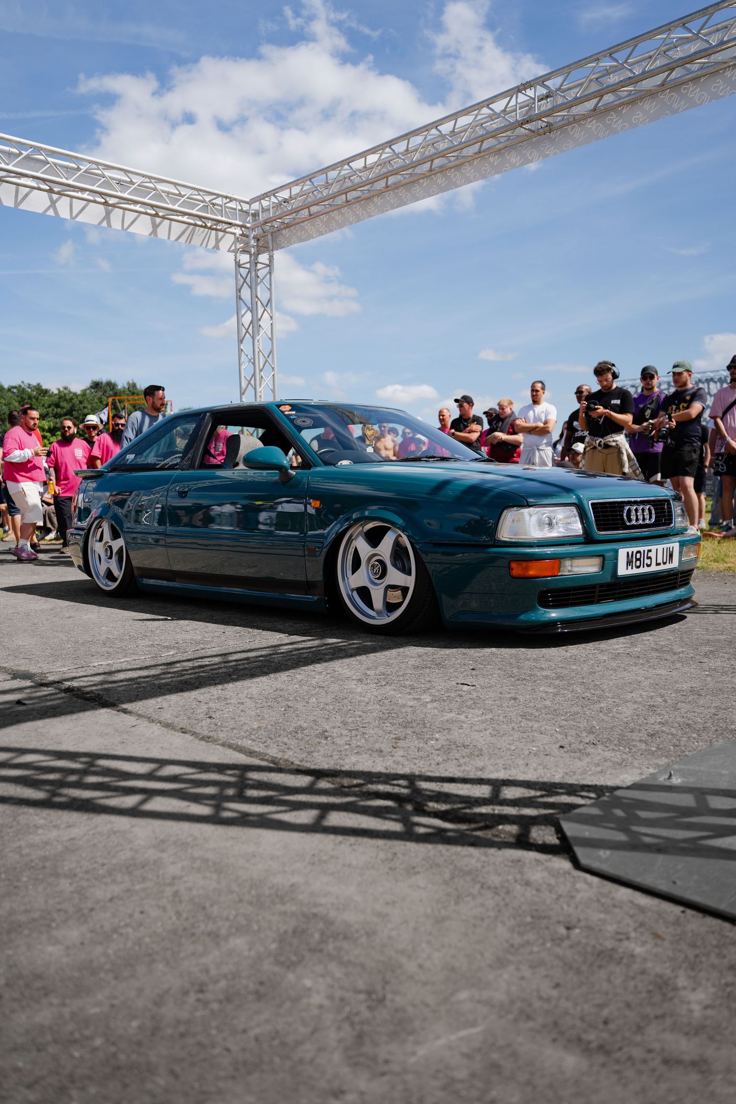 A lowered teal Audi RS2 on white wheels at a car show event with spectators watching under a metal truss gate.
