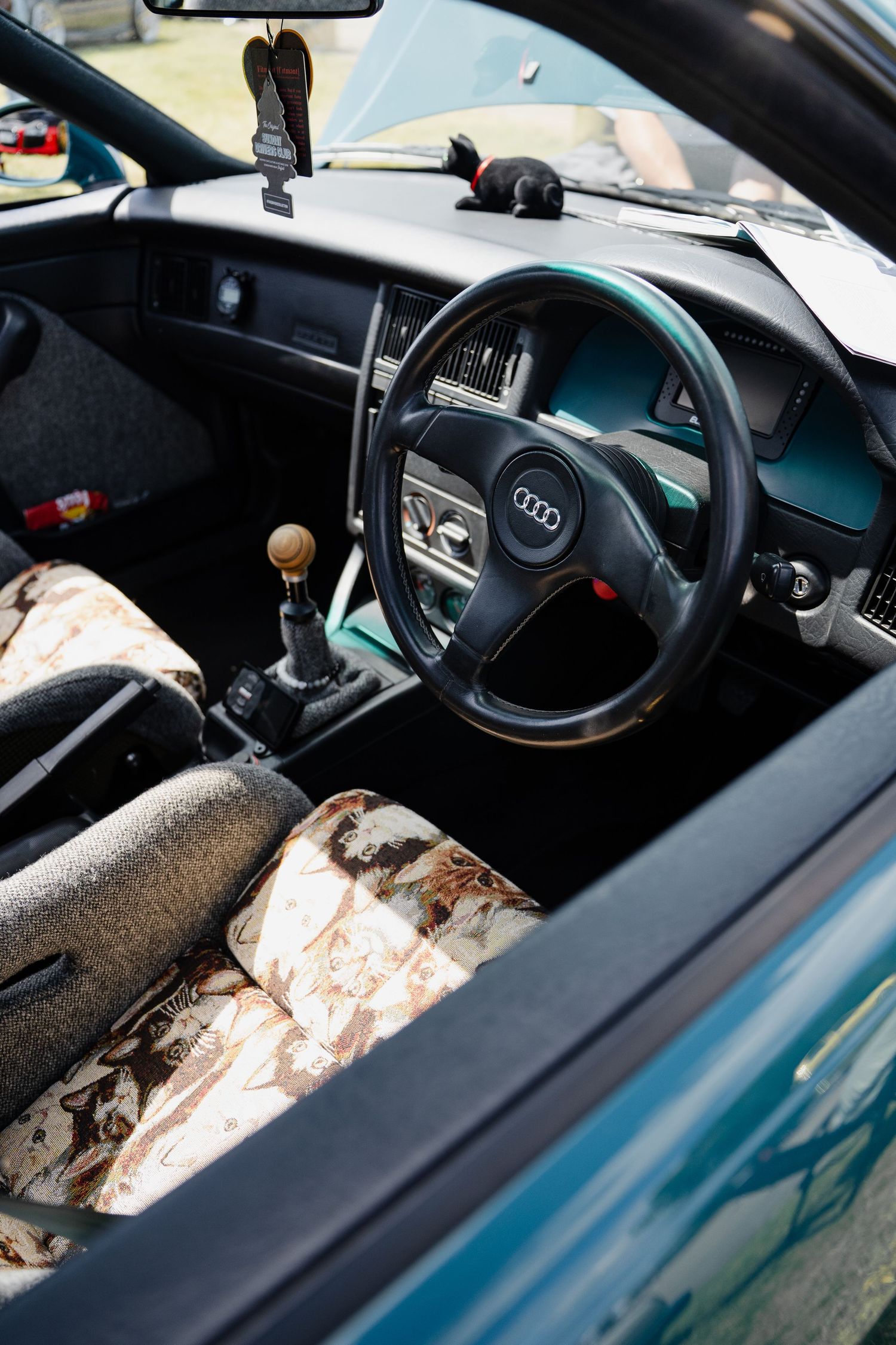 Vintage Audi car interior with worn floral seats, manual gearshift, and classic steering wheel with four-ring logo.