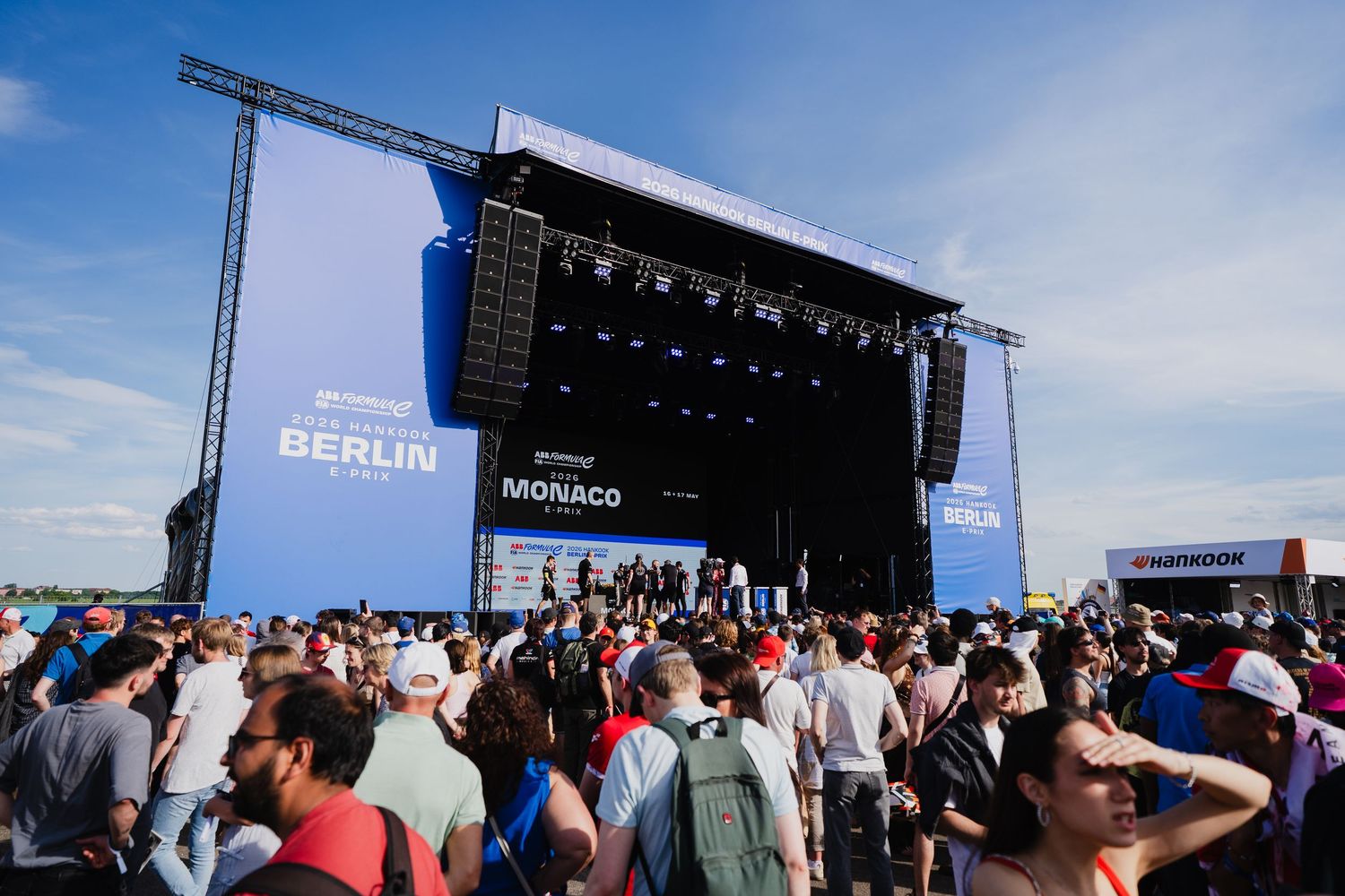 Large outdoor concert stage at Formula E Berlin and Monaco events with crowds of spectators under a clear blue sky.