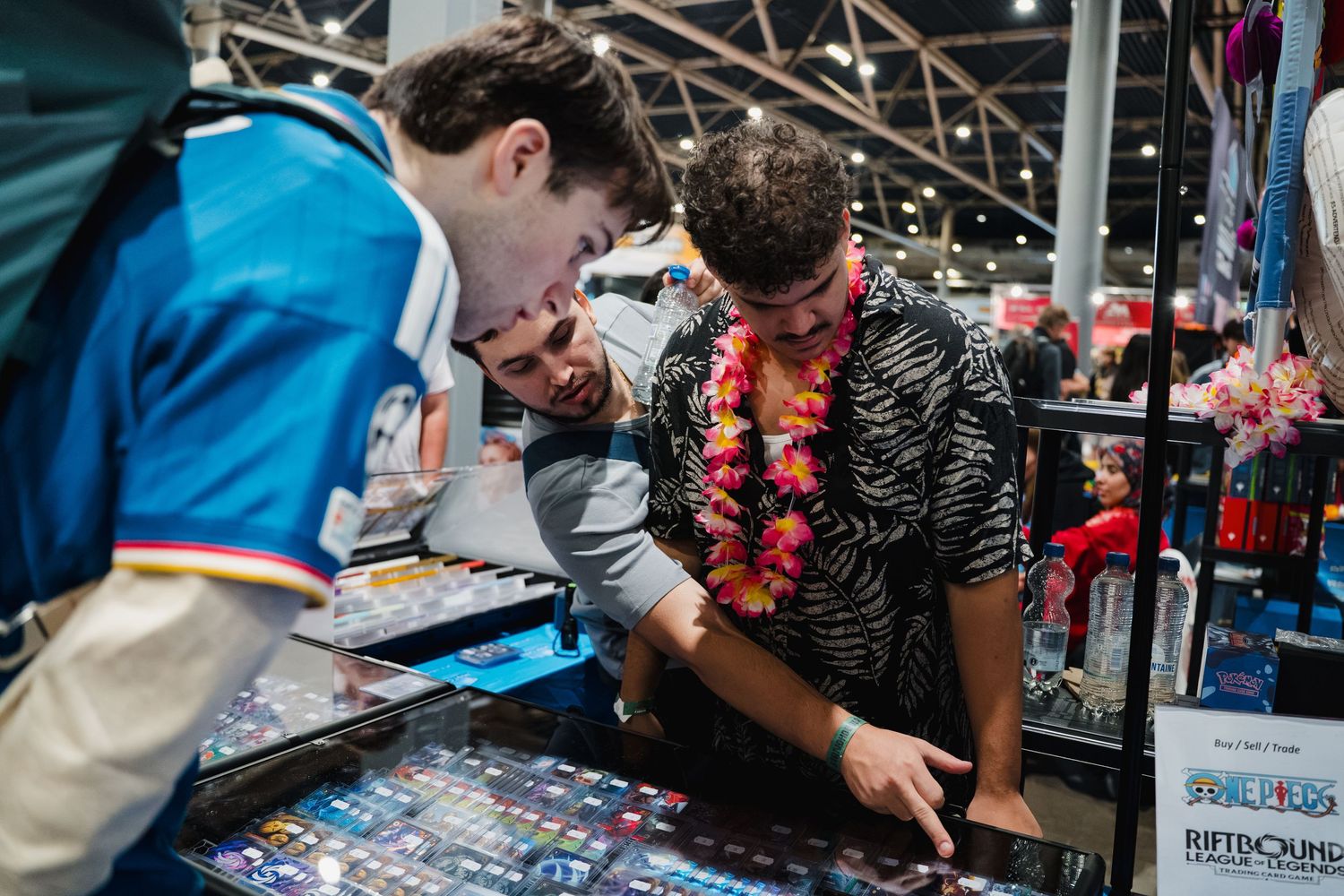 Three people examining trading cards at a convention booth with One Piece merchandise displays in the background.