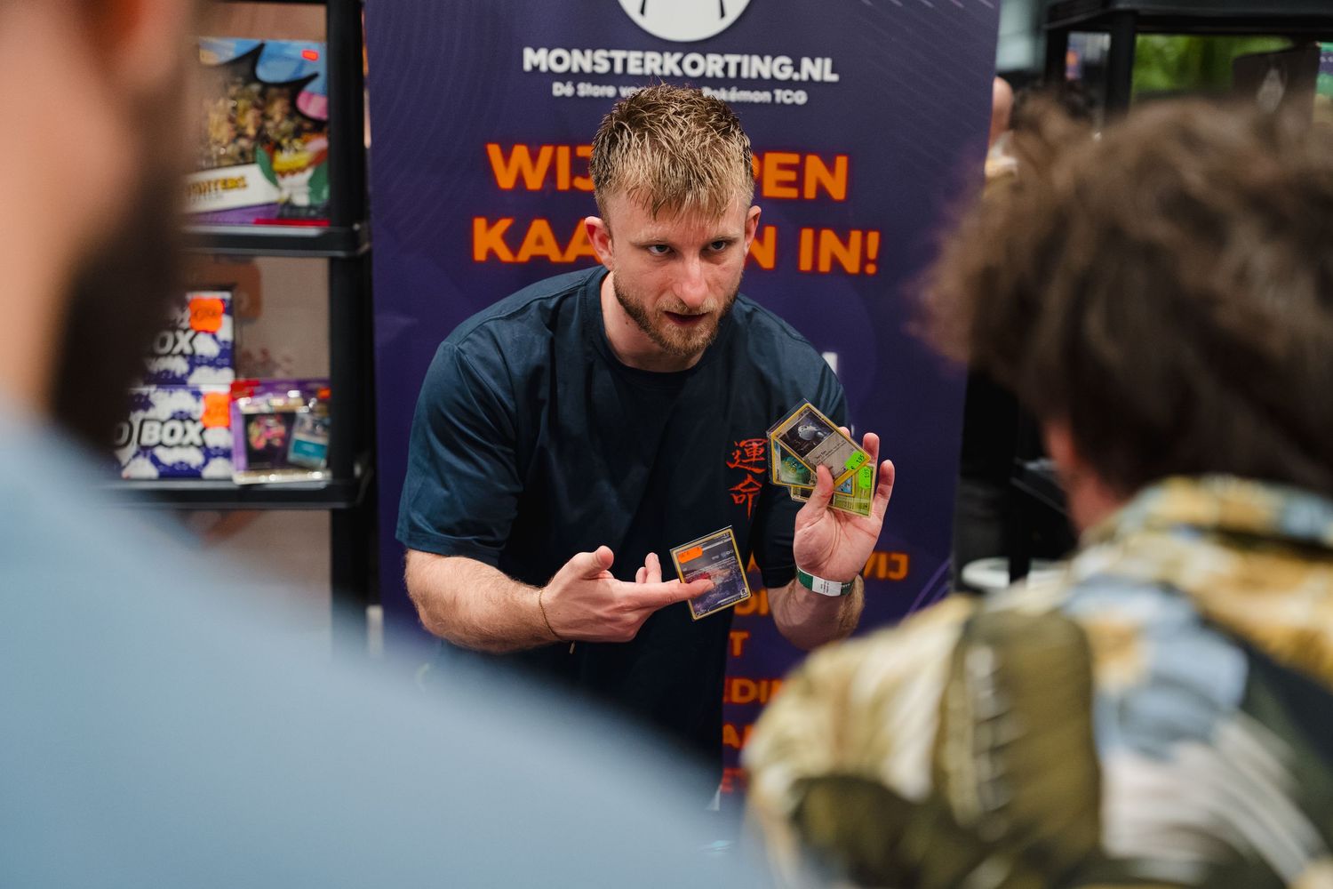 Man holding trading cards at Monsterkorting.nl booth, engaging with visitors at an indoor event.