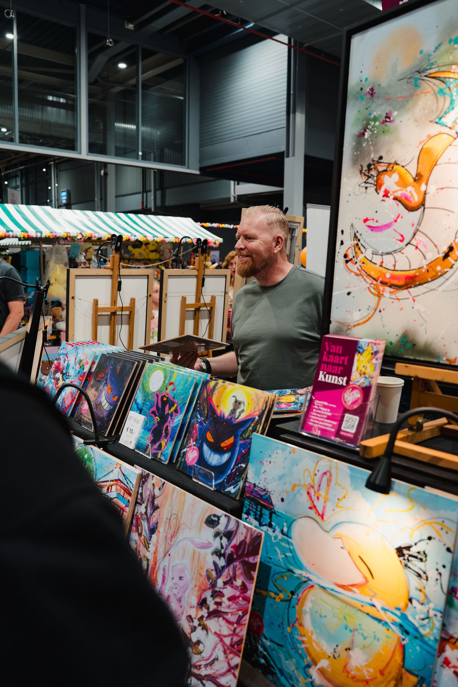 Red-haired man smiling at colorful art booth displaying vibrant paintings at an indoor art market.