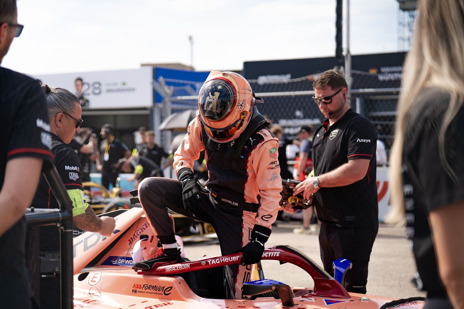 A race car driver in an orange helmet and suit climbs into a Formula car assisted by pit crew members at a racing event.