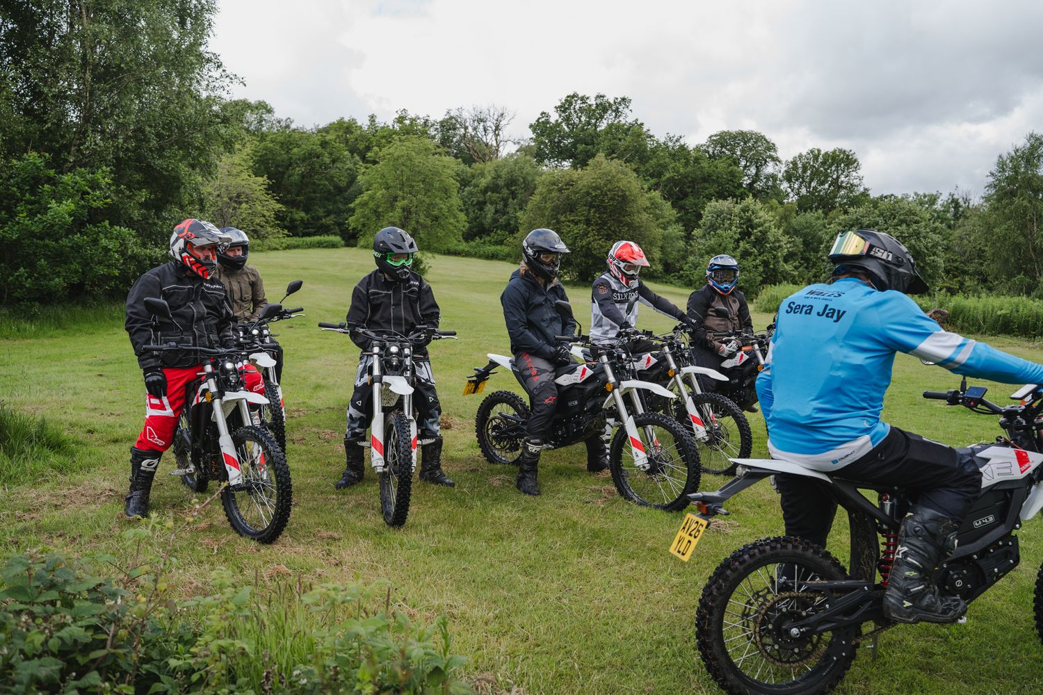 Group of dirt bike riders on electric motorcycles gathered in a green field listening to an instructor.