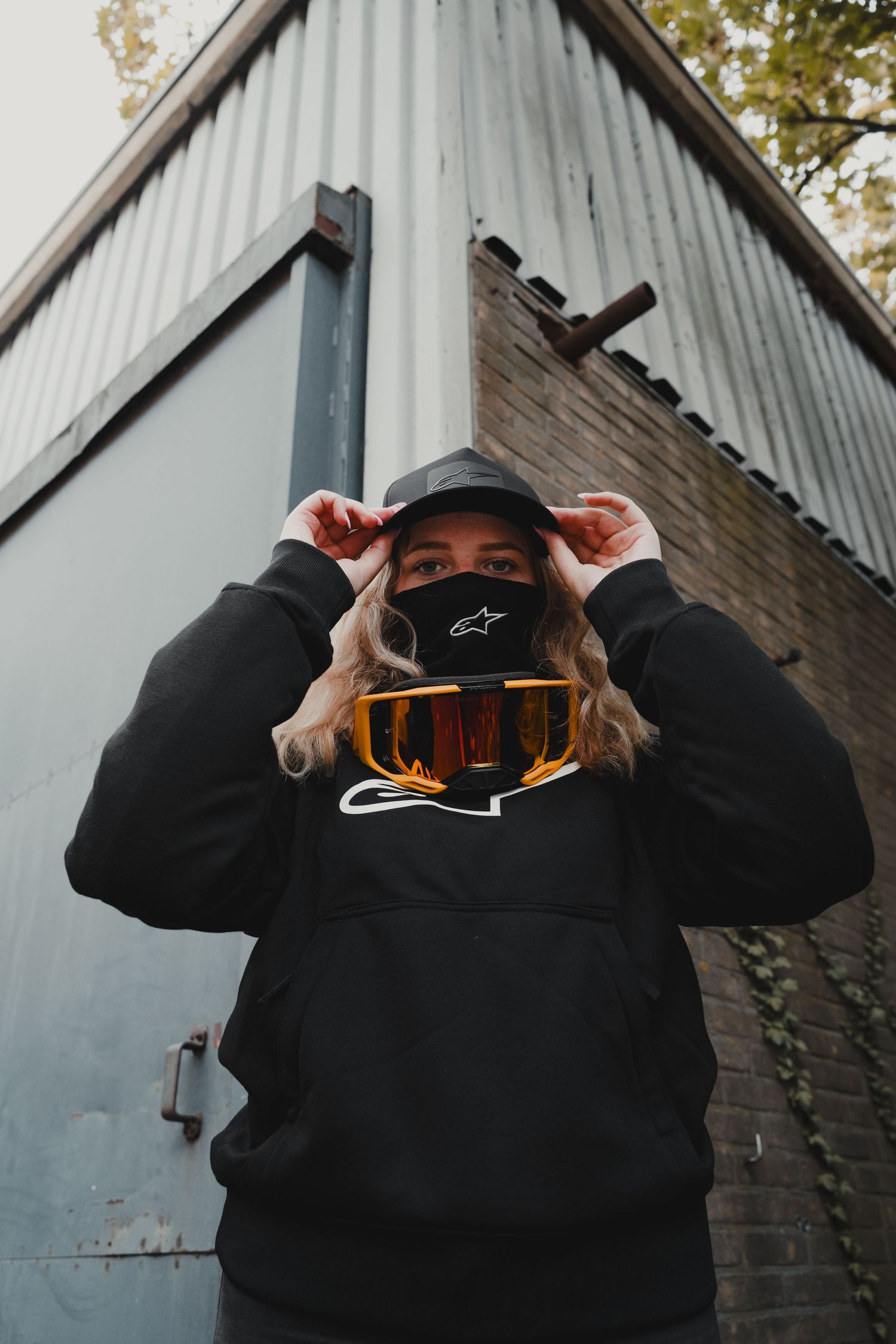 Person wearing black Alpinestars hoodie and cap with motocross goggles, posing outdoors near a metal building.