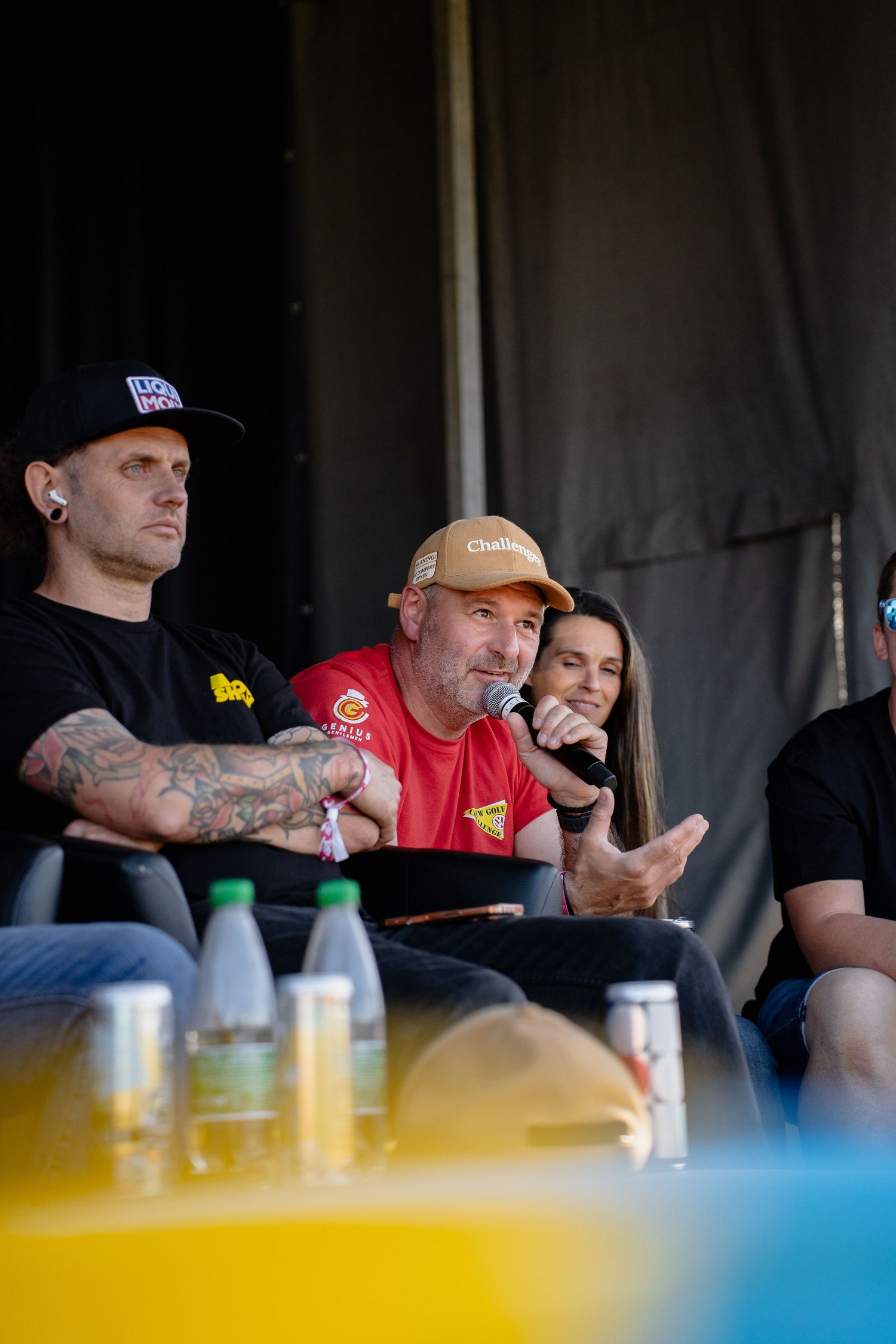 Three people seated at an outdoor panel event, man in red shirt speaking into microphone wearing gold cap.