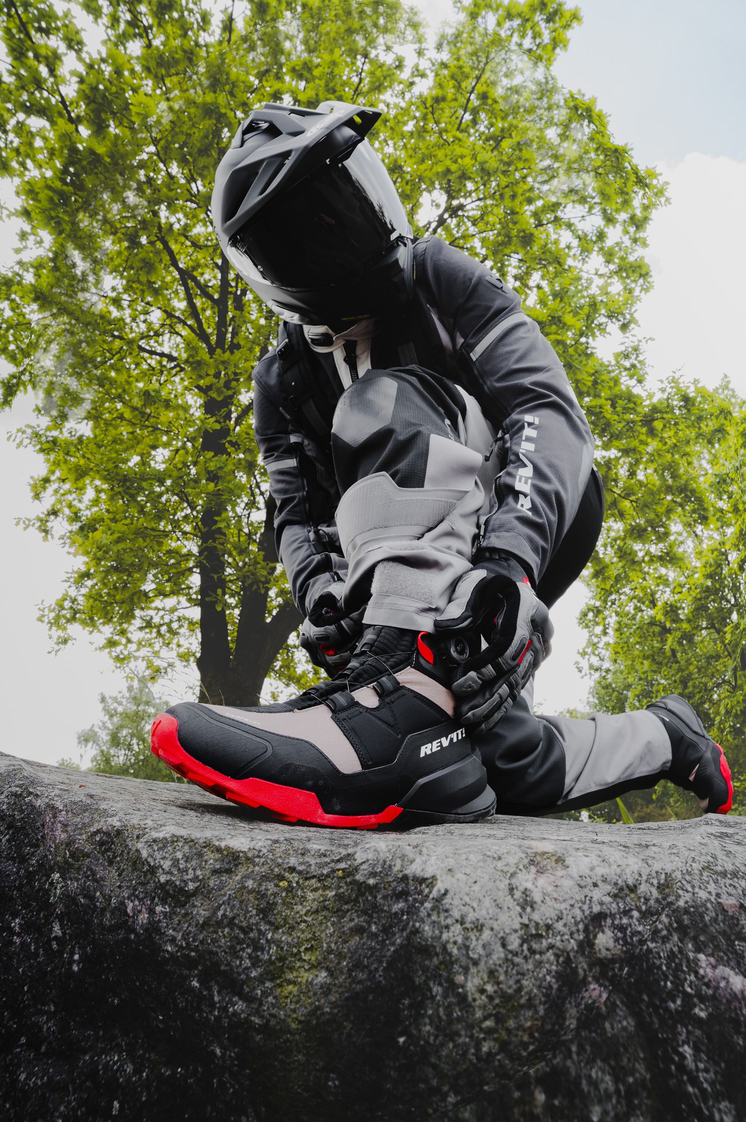 Motocross rider in full gear with red and black boots kneeling on a rock outdoors.
