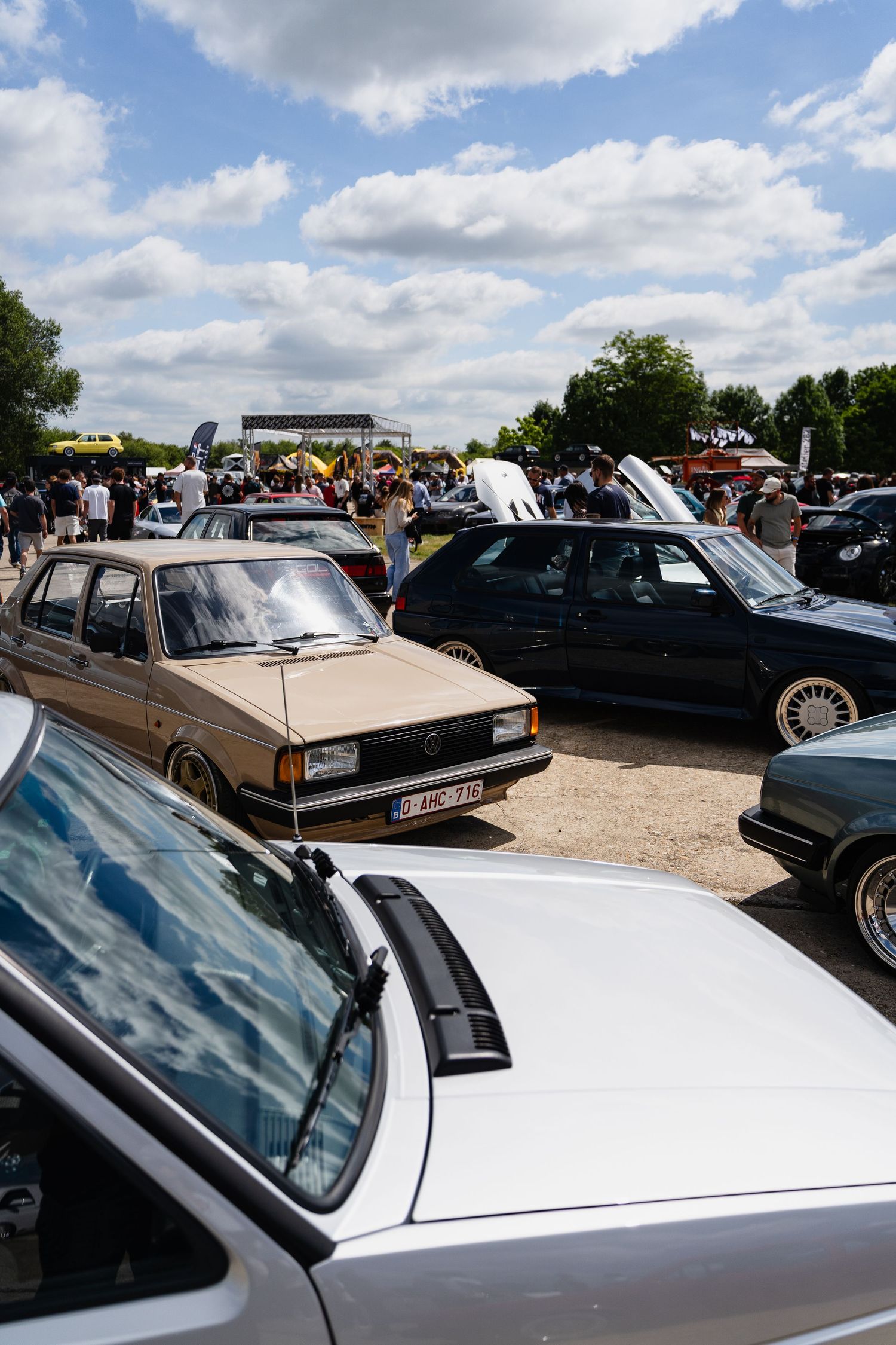 Vintage cars on display at an outdoor car show event under a partly cloudy sky with crowds of enthusiasts.