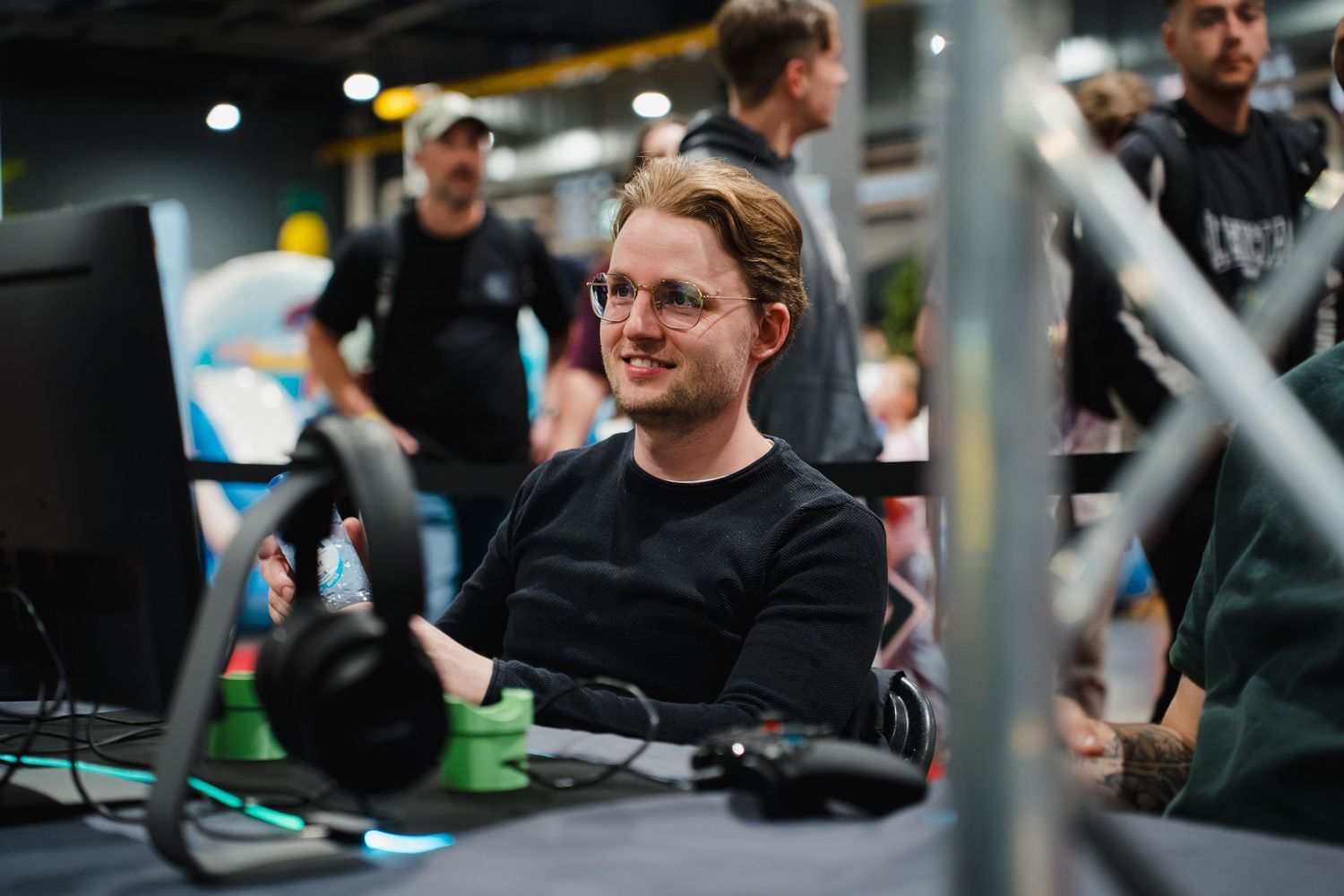 A smiling young man with glasses sits at a gaming setup with headphones and a monitor at an esports event.