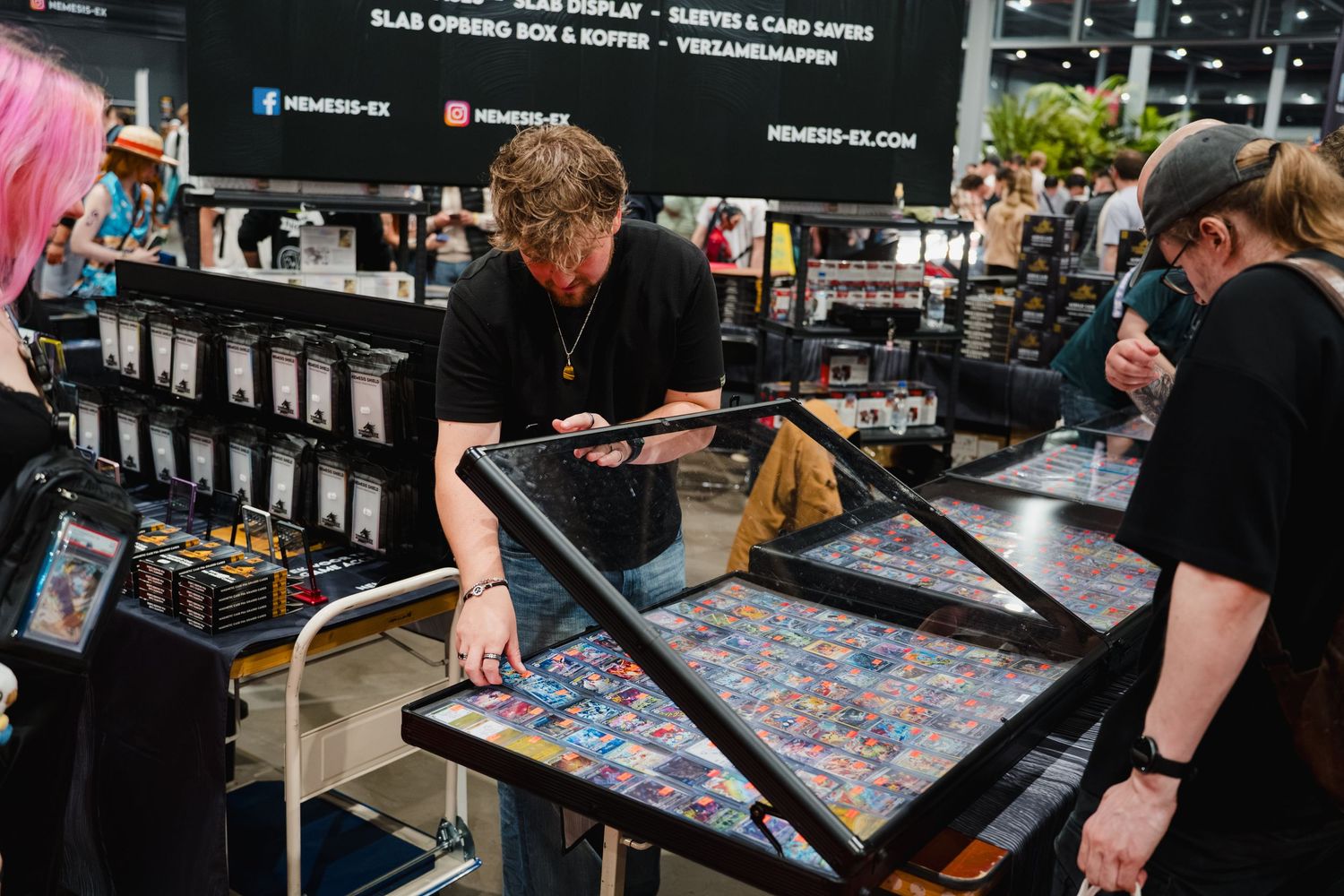 People browsing trading cards at a convention vendor booth with display cases and shelving units in the background.