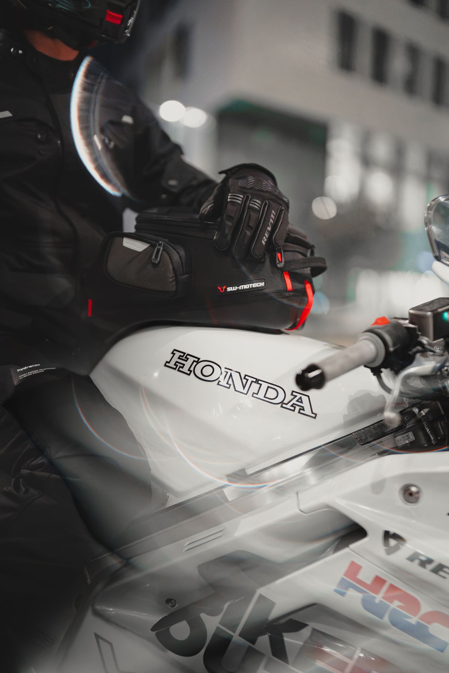 Motorcyclist in black gear riding a white Honda motorcycle, wearing a helmet and gloves in an urban setting.