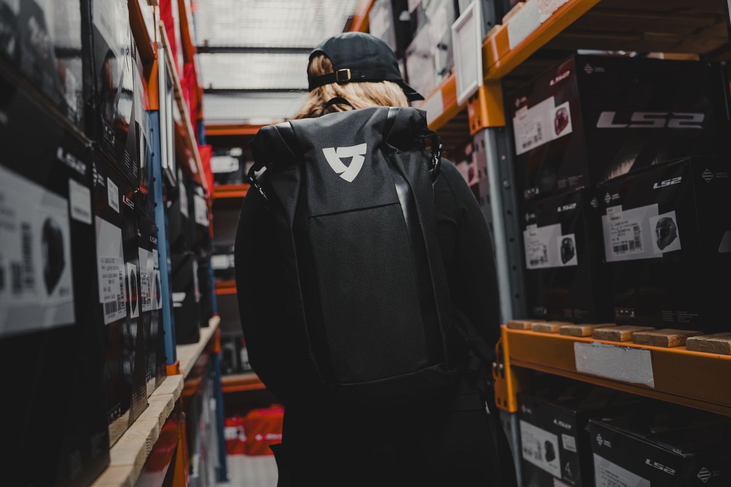 Person wearing a black backpack browsing shelves of helmets and gear boxes in a motorcycle equipment warehouse.