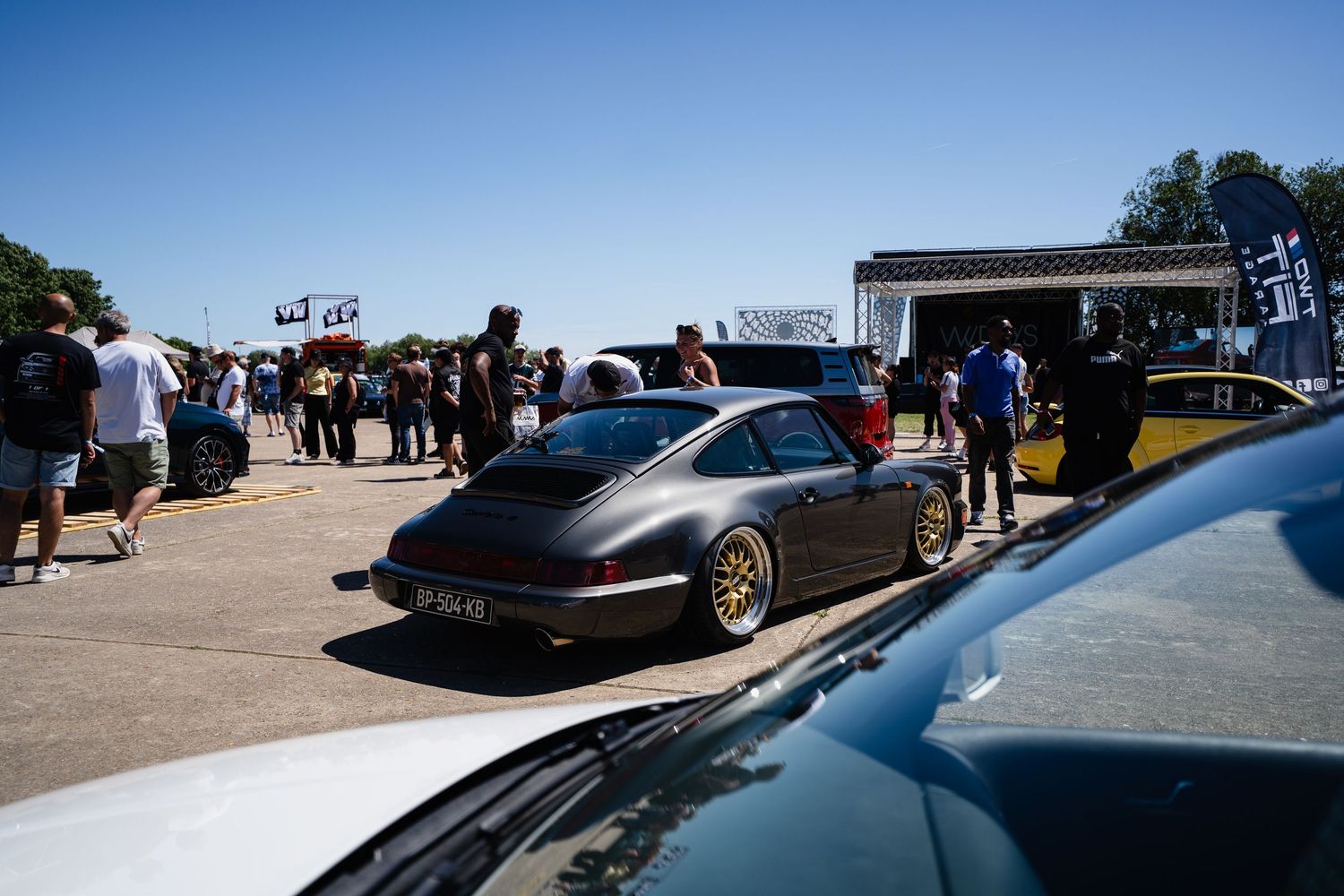 Dark Porsche 911 with gold wheels at an outdoor car show event on a sunny day.