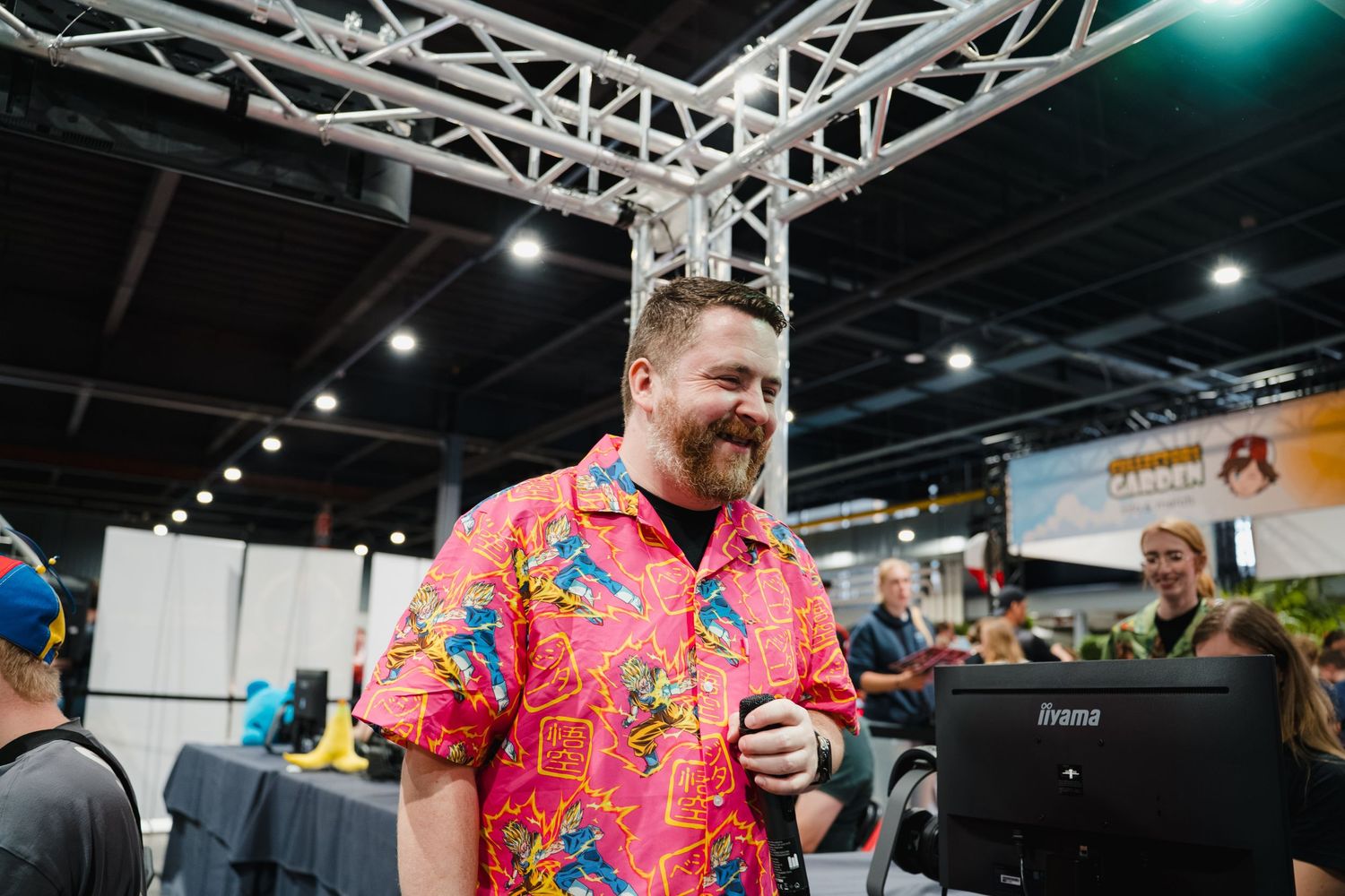 Man in colorful Hawaiian shirt smiling at a gaming convention booth with monitors and attendees in background.