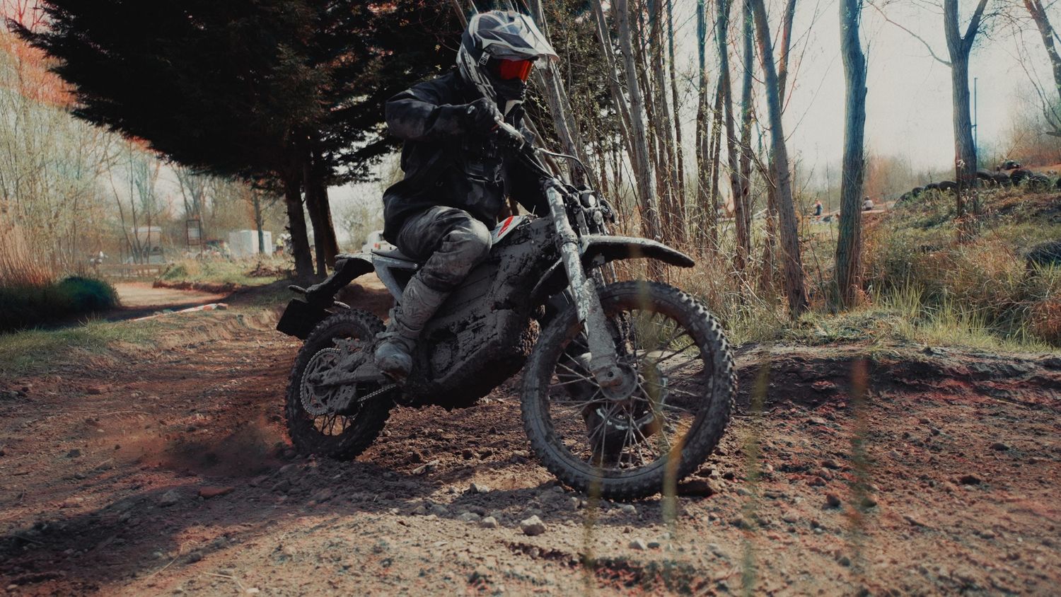 A muddy dirt bike rider navigates rough terrain on a motocross trail surrounded by trees.