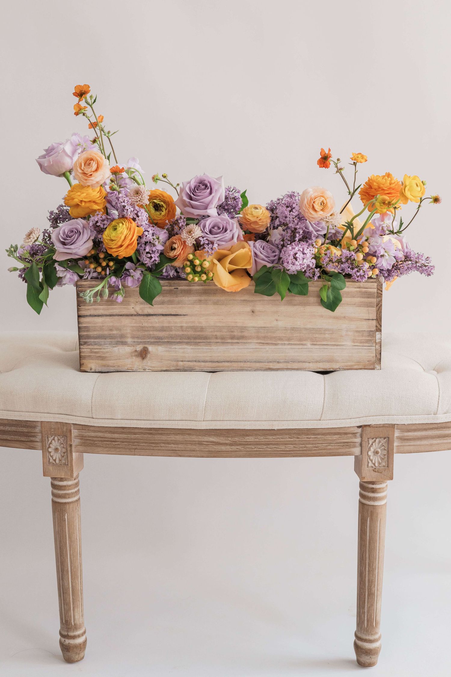 Planter box arrangement with ranunculus lilac roses geum stock flower hypercium orange and purple on tufted bench