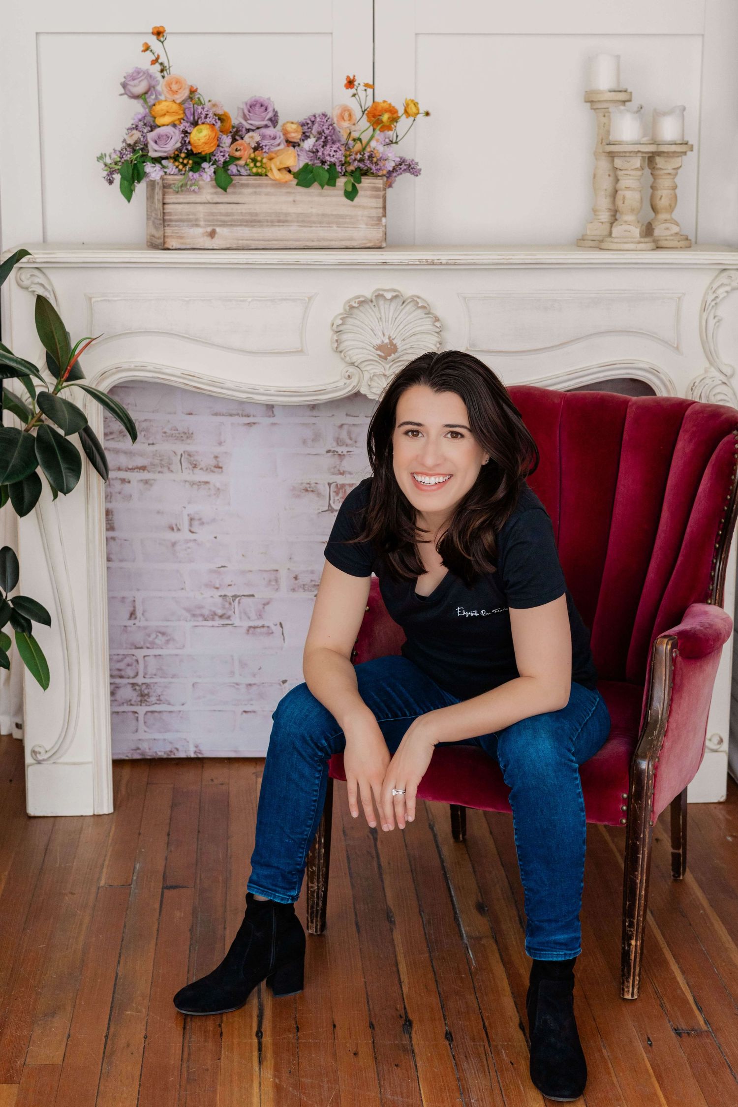 woman sitting in a rouge armchair while smiling in front of faux fireplace with planter box arrangement on mantel