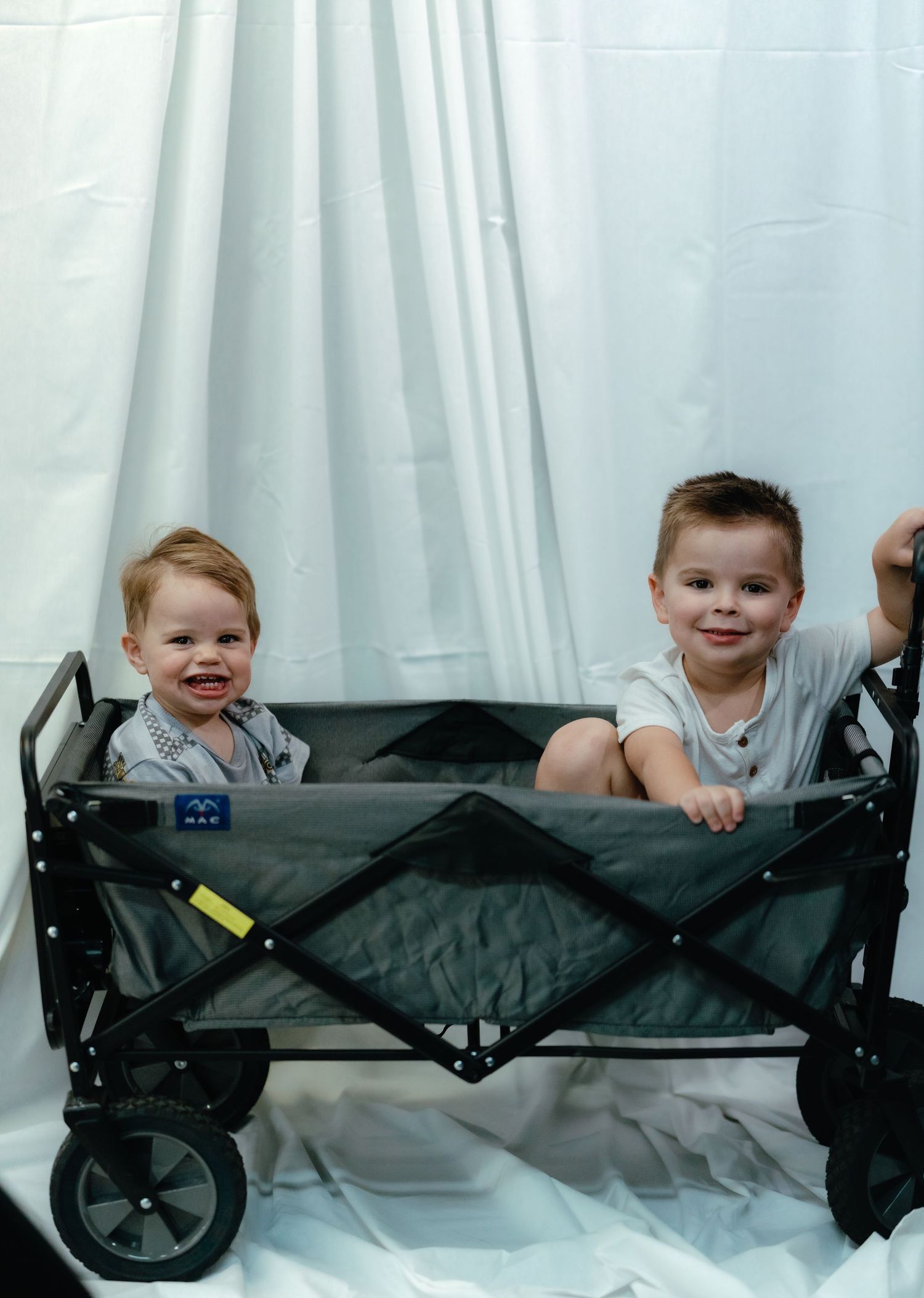 Baby plays with folded portable wagon against white backdrop.