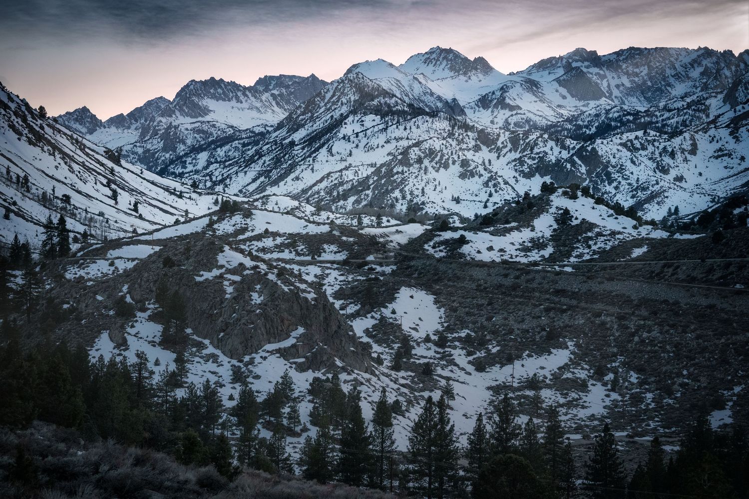 Snow-covered mountain peaks and pine trees at dusk in a dramatic alpine valley landscape.