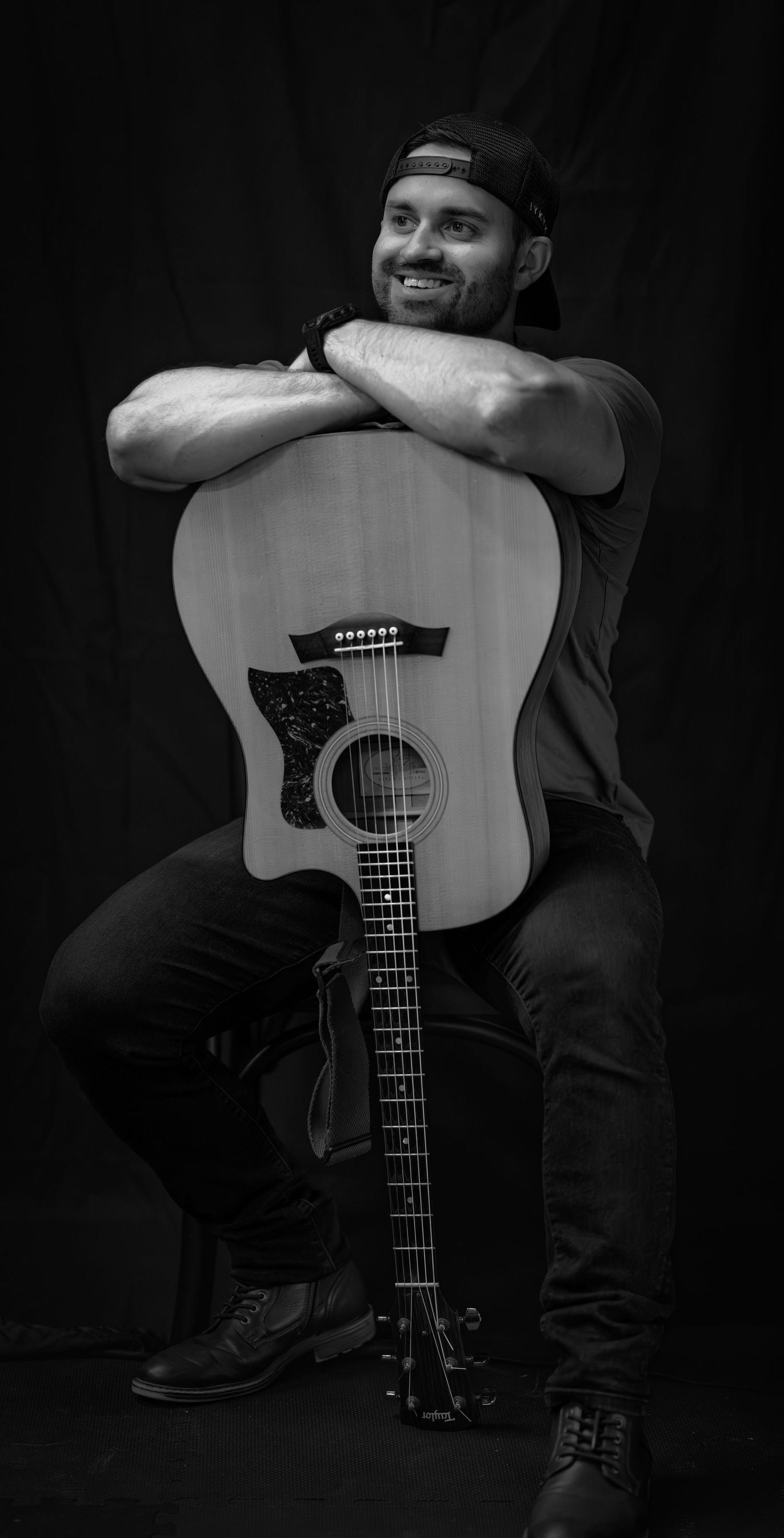 Black and white portrait of a musician posing with an acoustic guitar against a dark background.