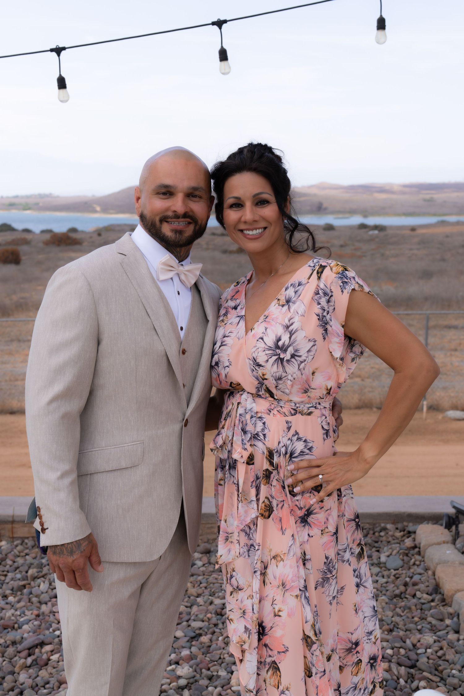 Couple poses for photos at desert venue with string lights and palm trees in background.