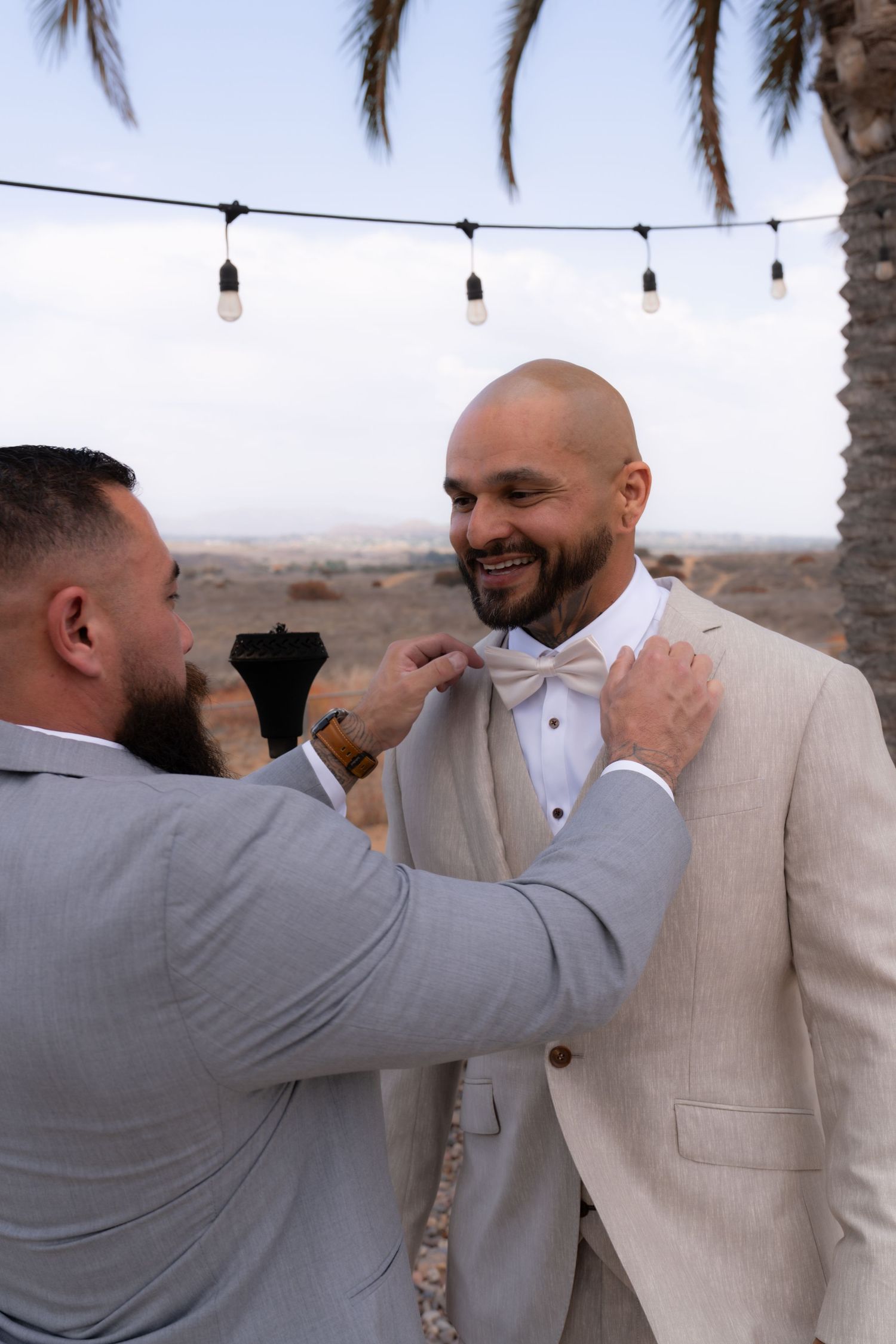 Two men in formal suits share an emotional moment at an outdoor desert venue with string lights.