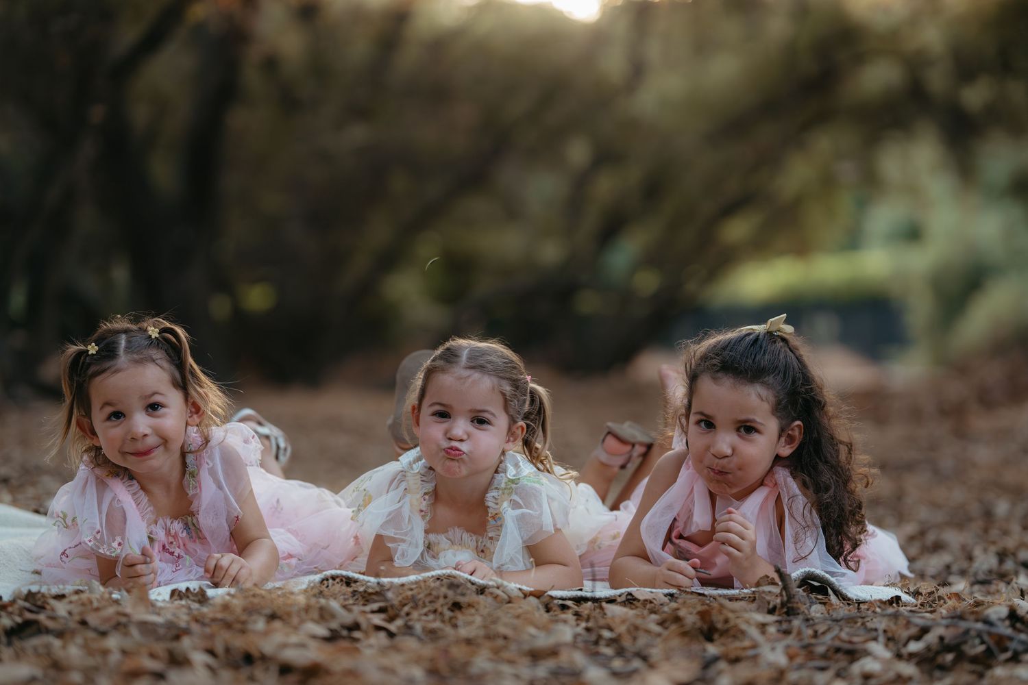 A series of young girls in white dresses lying on their stomachs in a row on autumn leaves during a photo shoot.