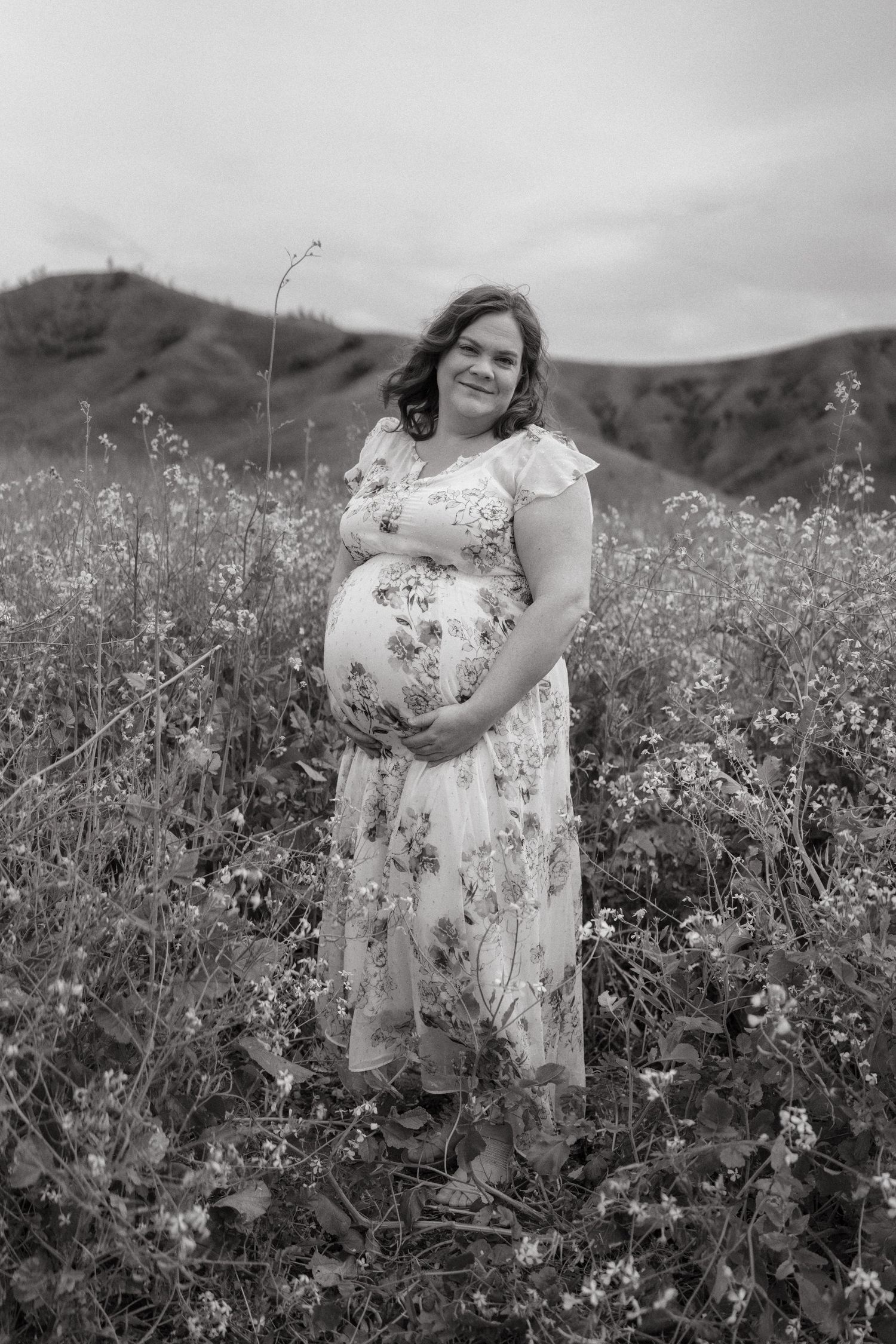 A person in a floral dress poses thoughtfully in a blooming wildflower field on an overcast day.