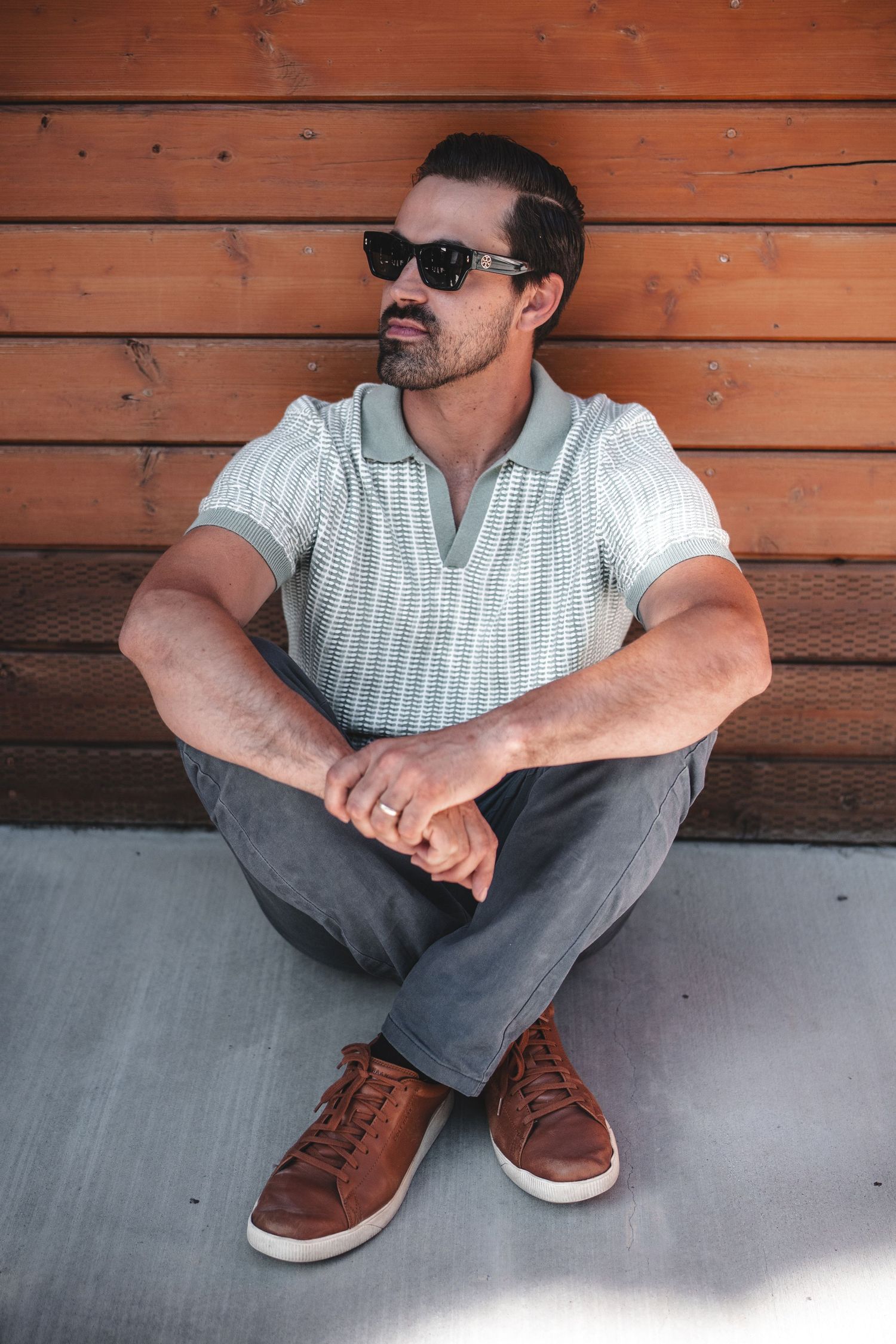 Person in casual attire and sunglasses sits against wooden wall in natural light.