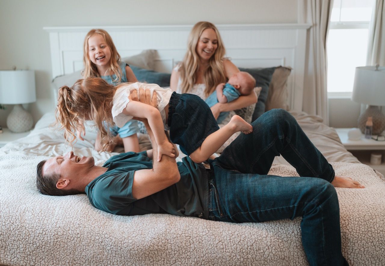 Parent and child lay together on a bed while playing and laughing in a bright sunlit room.