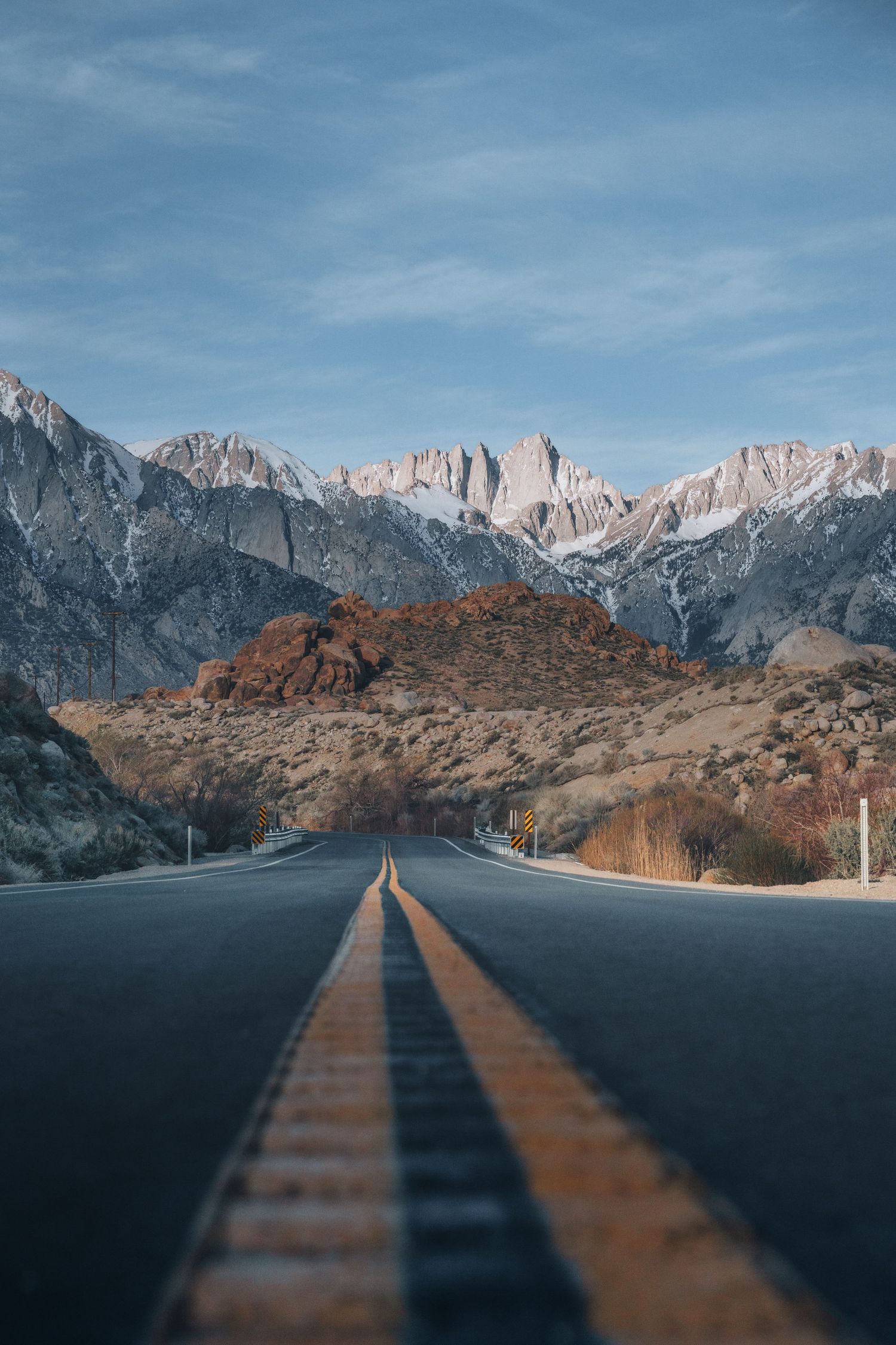 A straight desert road with yellow center lines leads toward snow-capped Sierra Nevada mountains under a blue sky.