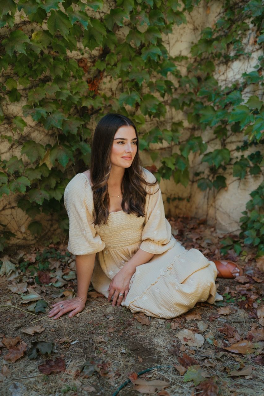 Woman in vintage cream dress poses elegantly against ivy-covered wall.