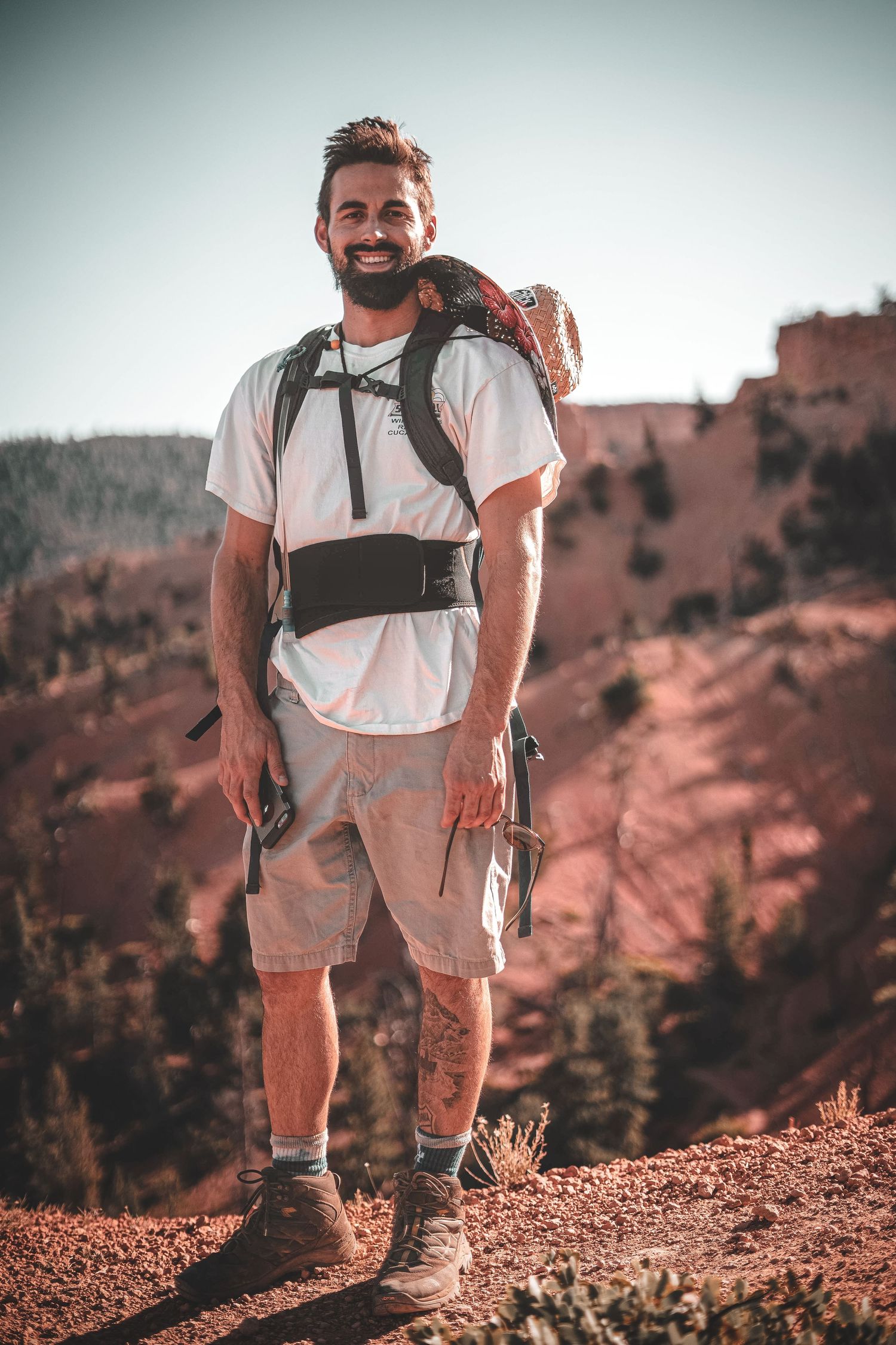 Hiker in white t-shirt and backpack stands on red rock formation in desert landscape.