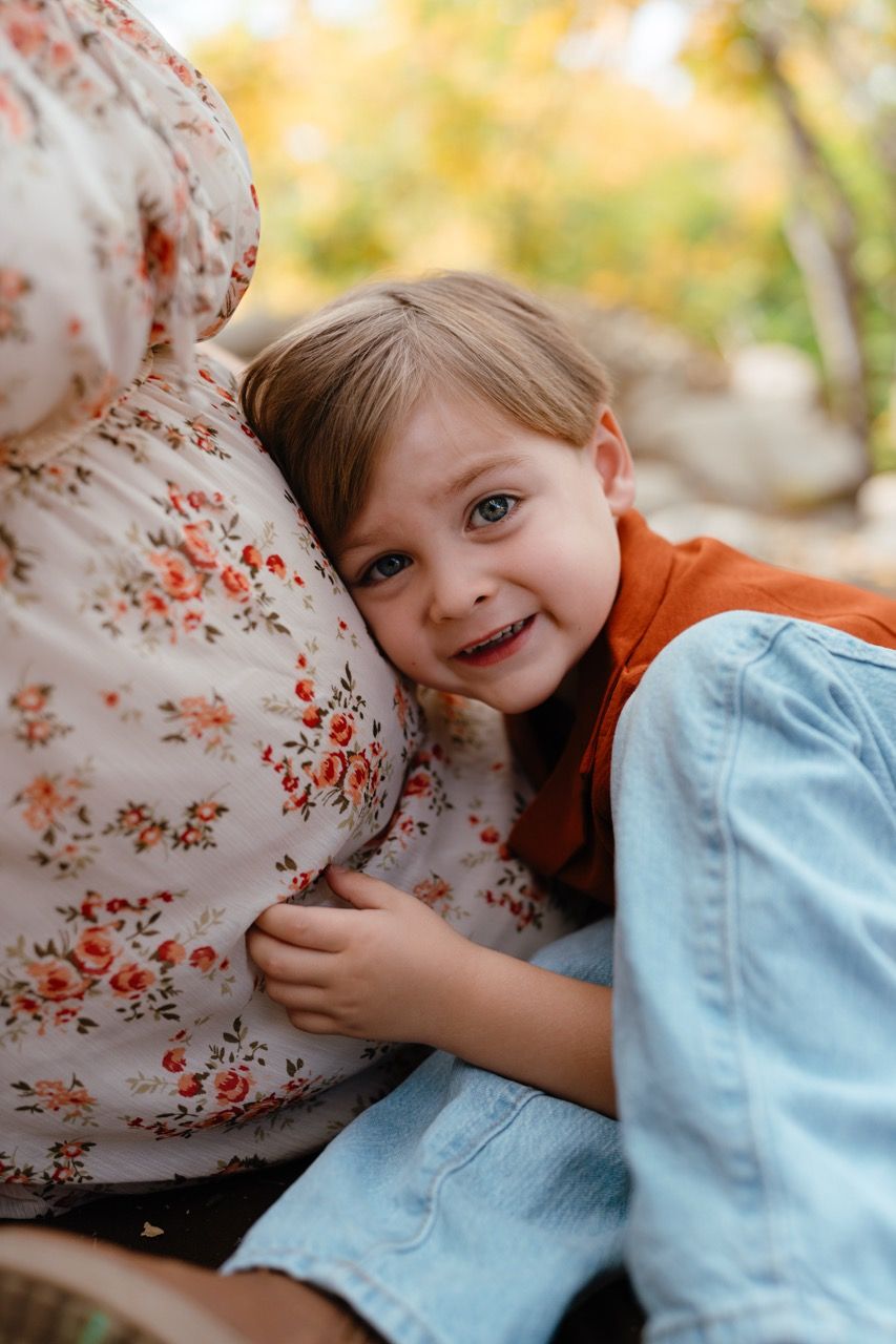 A young child in orange and denim hugs his mother's pregnant belly with autumn colors in the background.