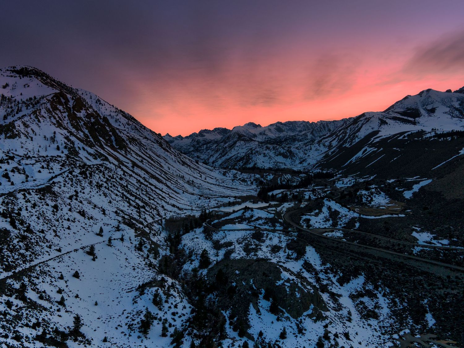 Aerial view of snow-covered mountain valley at sunset with vibrant pink and purple sky over winding road.