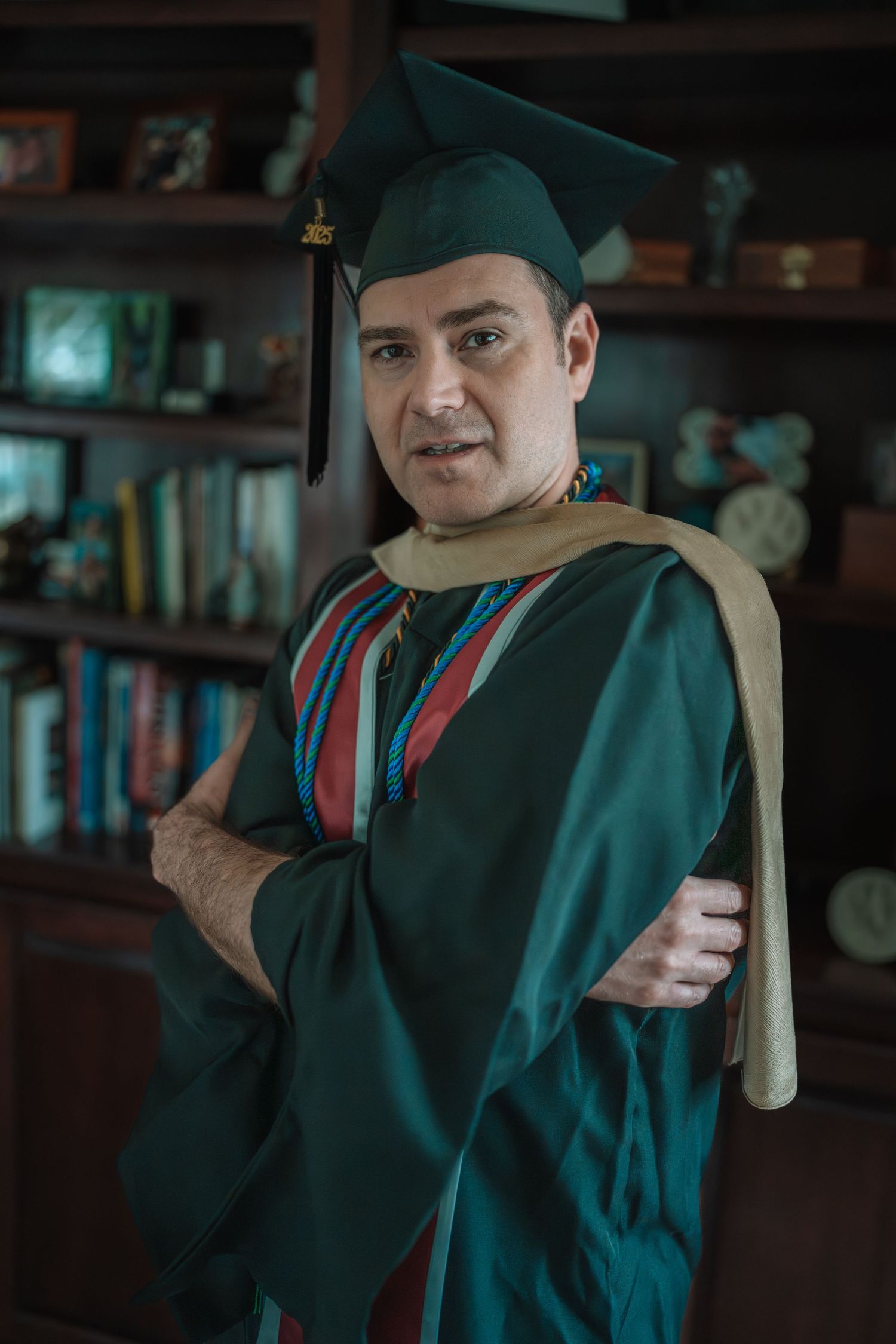 Graduate in dark green academic regalia poses with diploma in library setting.