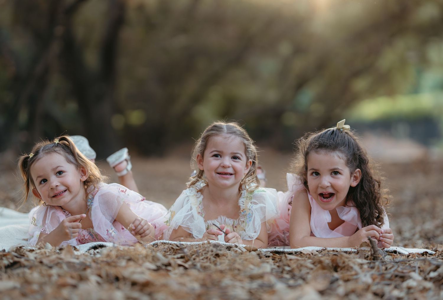 Three children in pink dresses smiling while lying on fallen leaves.