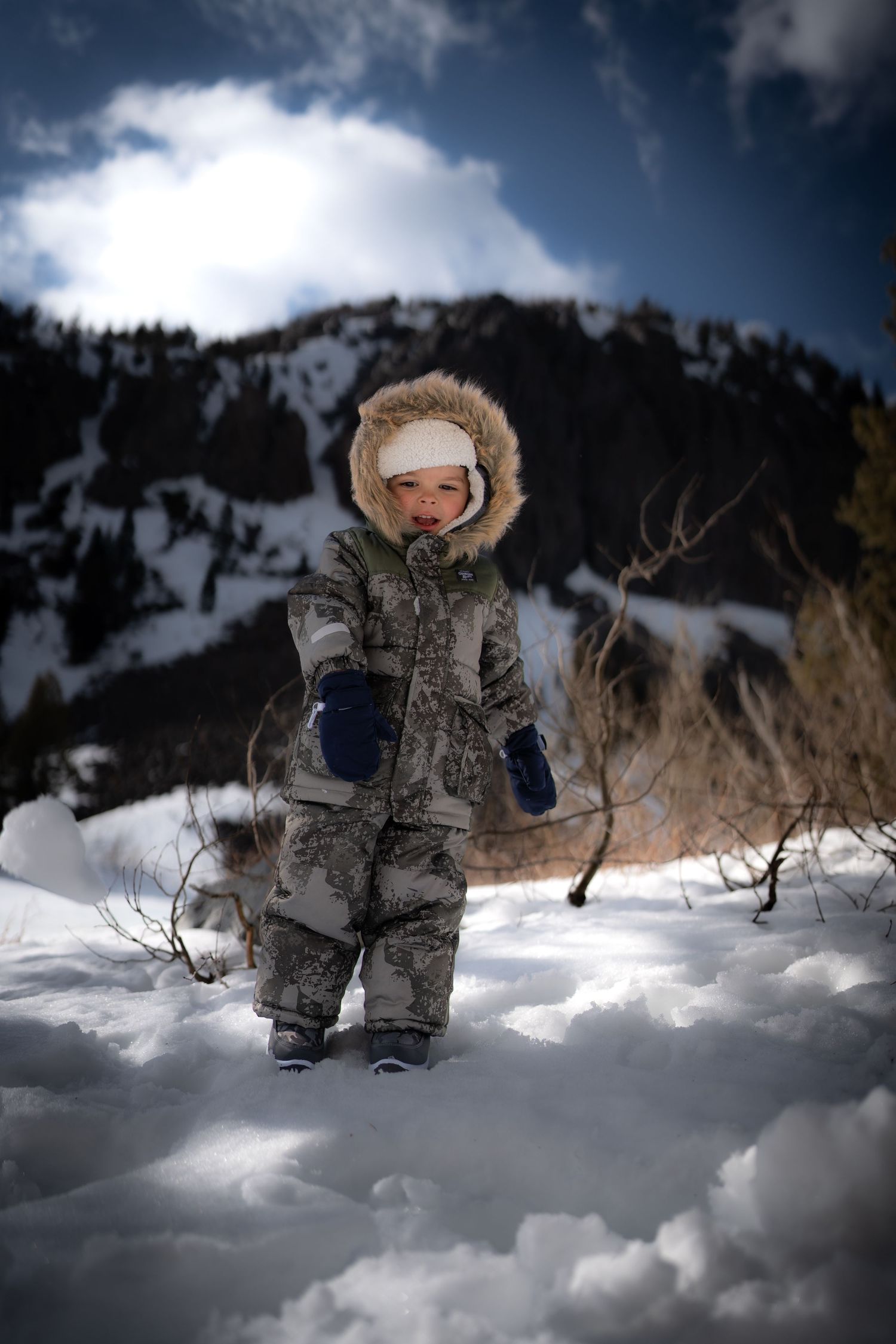 Small figure in winter snowsuit standing in deep snow with mountain backdrop.