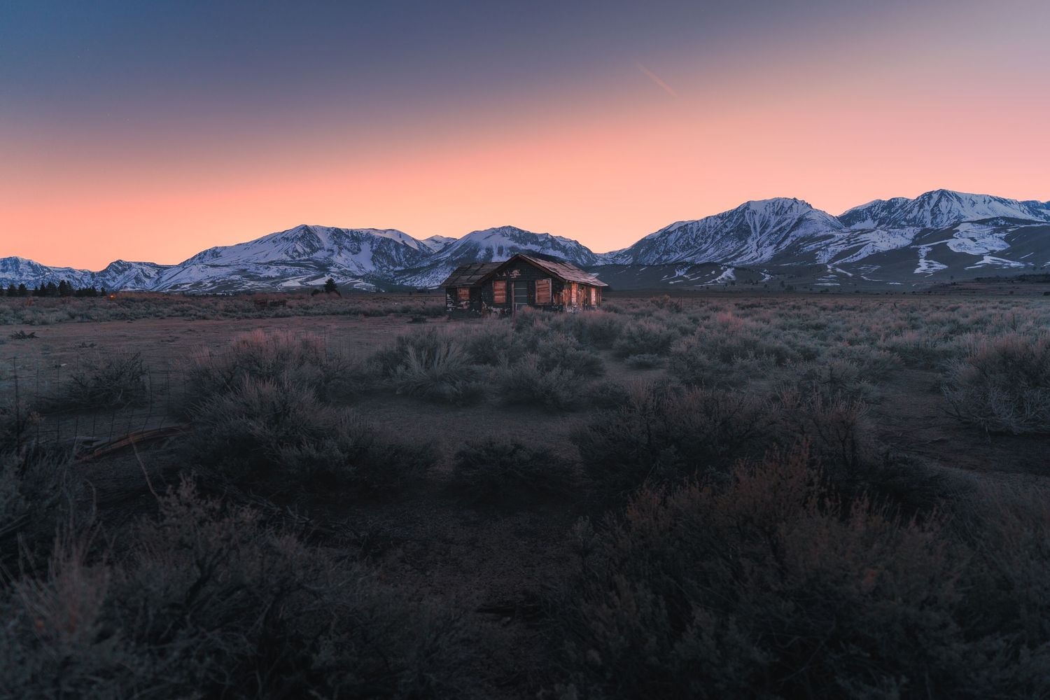 Abandoned rustic cabin in desert scrubland at sunset with snow-capped mountains and pink sky in background.