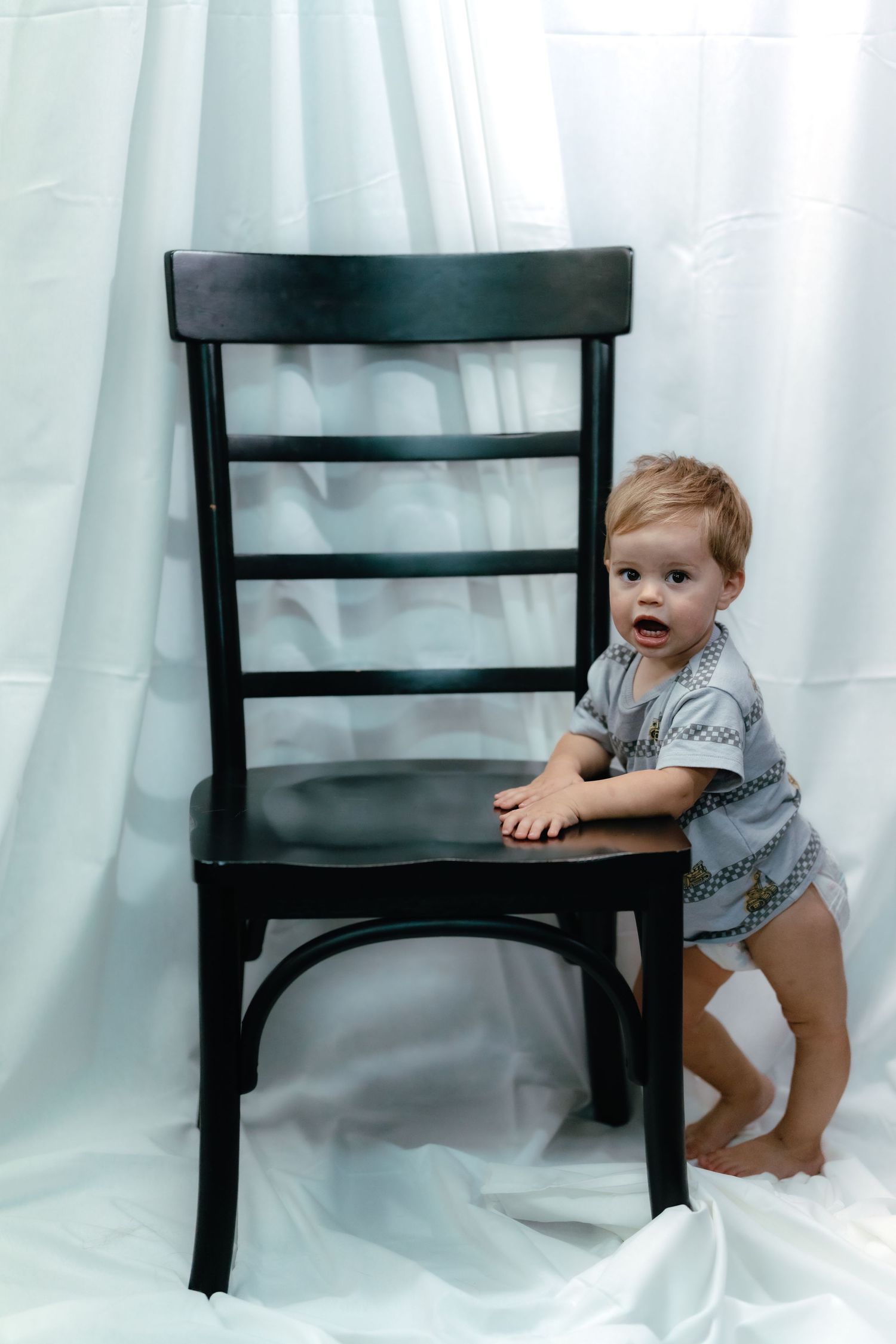 Small child plays and climbs on black wooden chair against white backdrop.