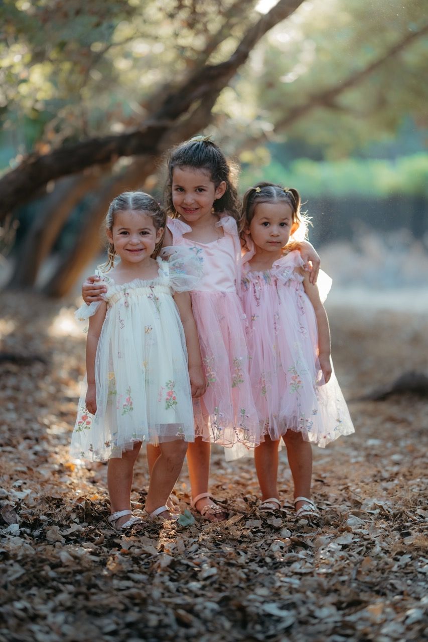 Three young children in pink and white dresses stand together on fallen autumn leaves in a park setting.
