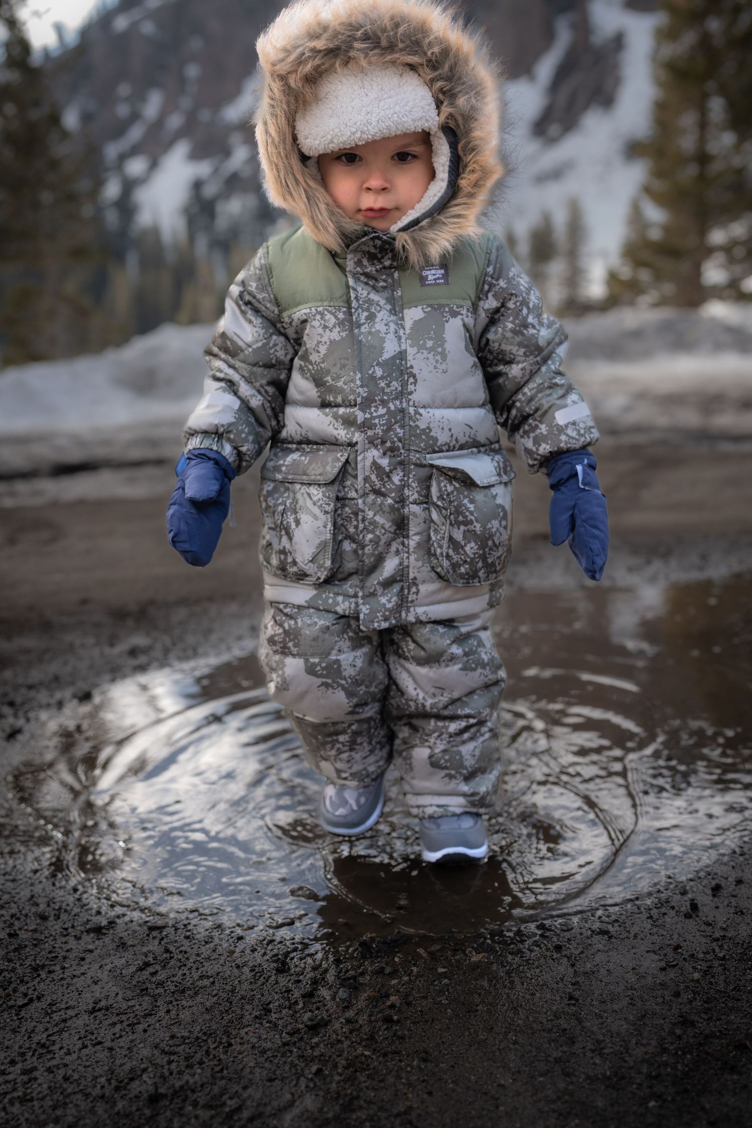 Small figure in winter gear standing in melted snow puddle with mountains behind.