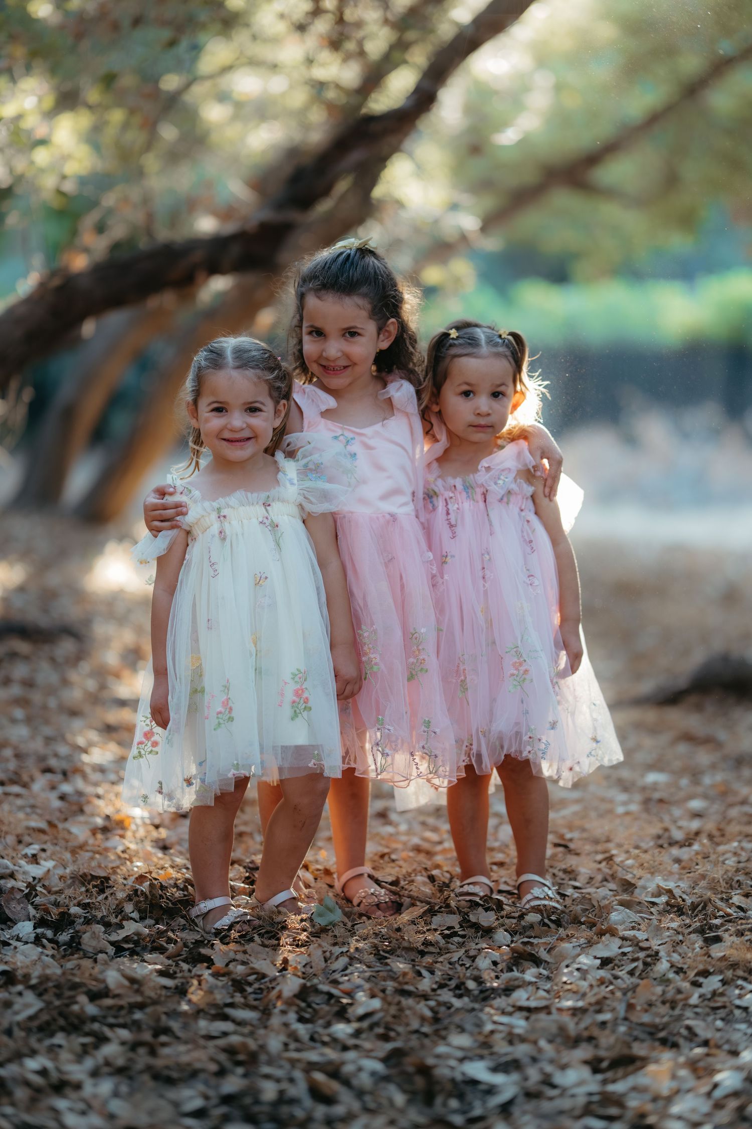 Three young children in pink and white dresses play together in fallen autumn leaves.