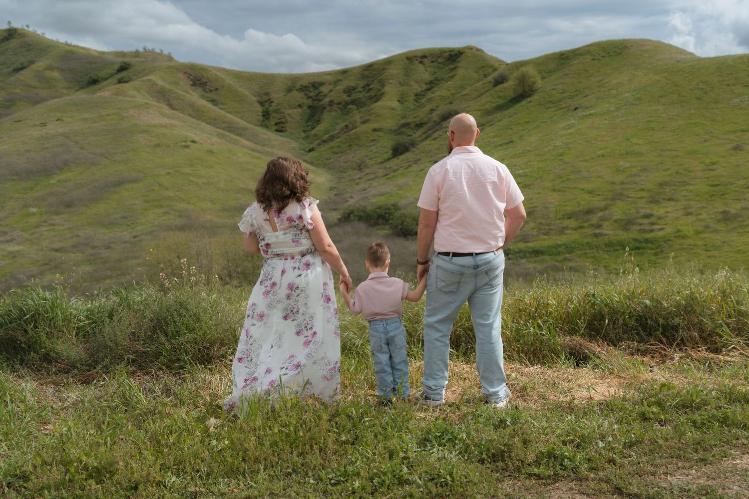 Three people sit together on a grassy hillside overlooking scenic rolling green mountains in the distance.