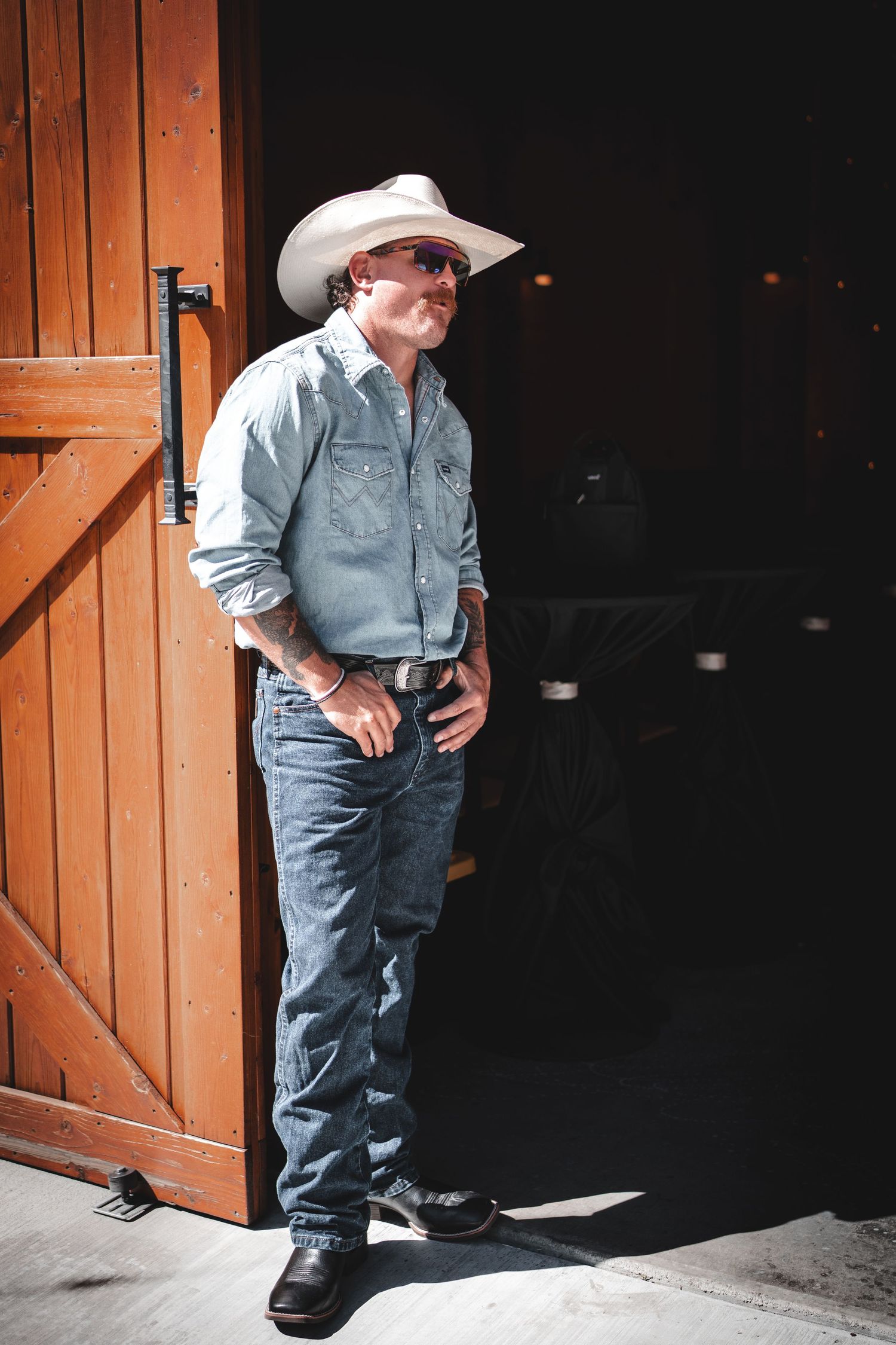 Person in cowboy attire and denim leans against wooden barn door in dramatic lighting.