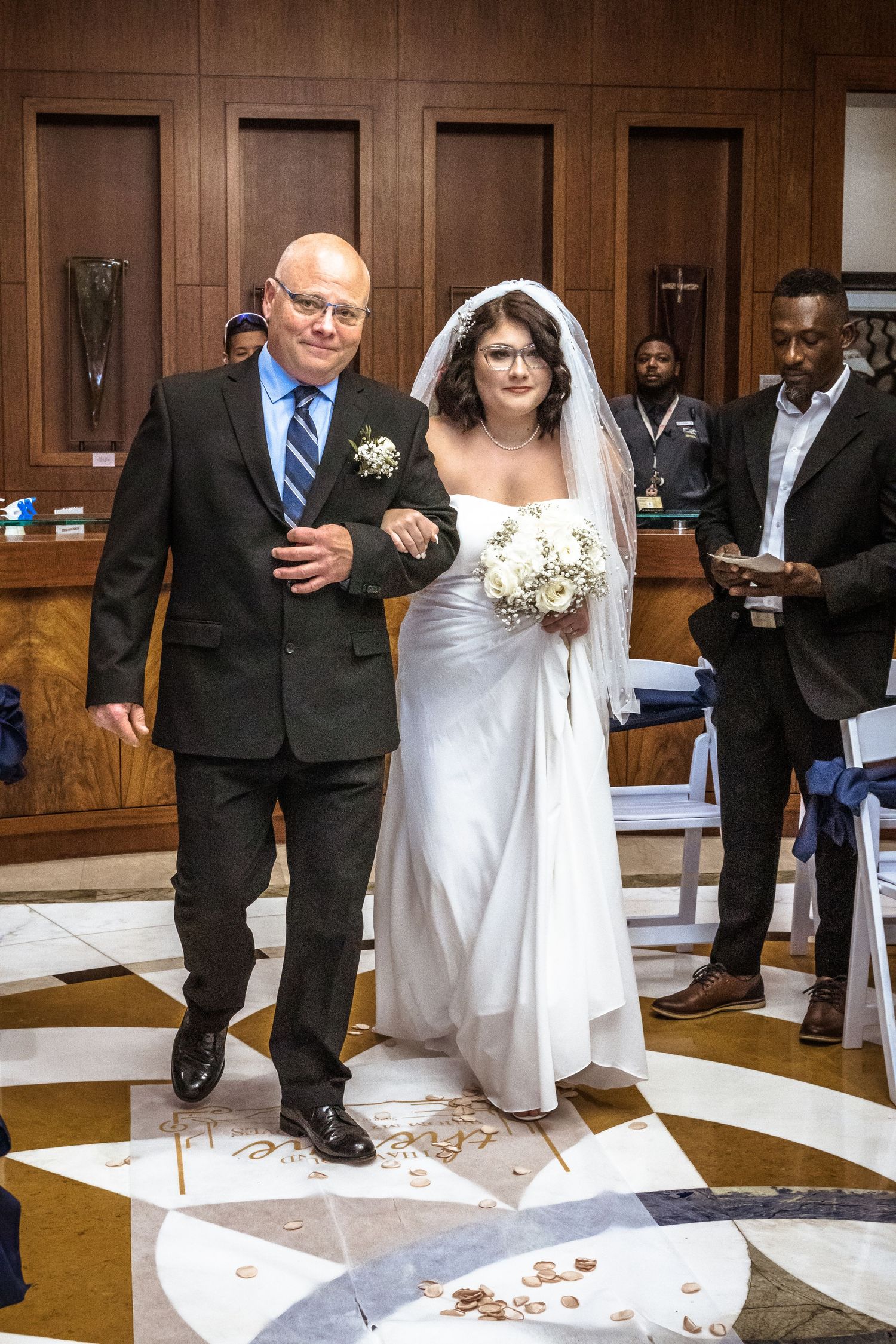 father walking bride down isle of wedding. father in black suite, bride in white. brown walls in background.